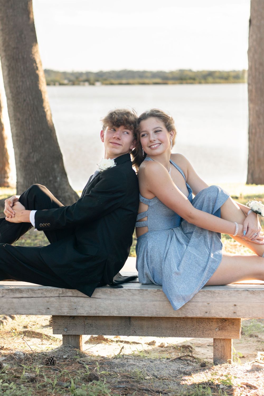 A couple sits back-to-back on a wooden bench by a lake.