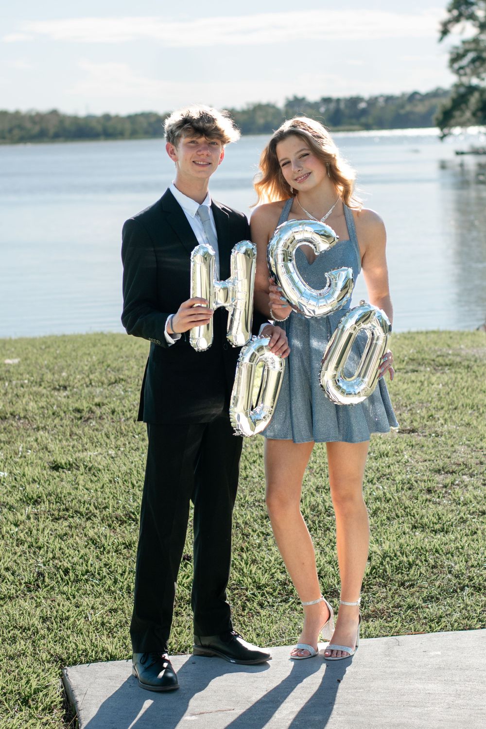 A couple holds silver balloon letters spelling 'HOCO' by a lake.