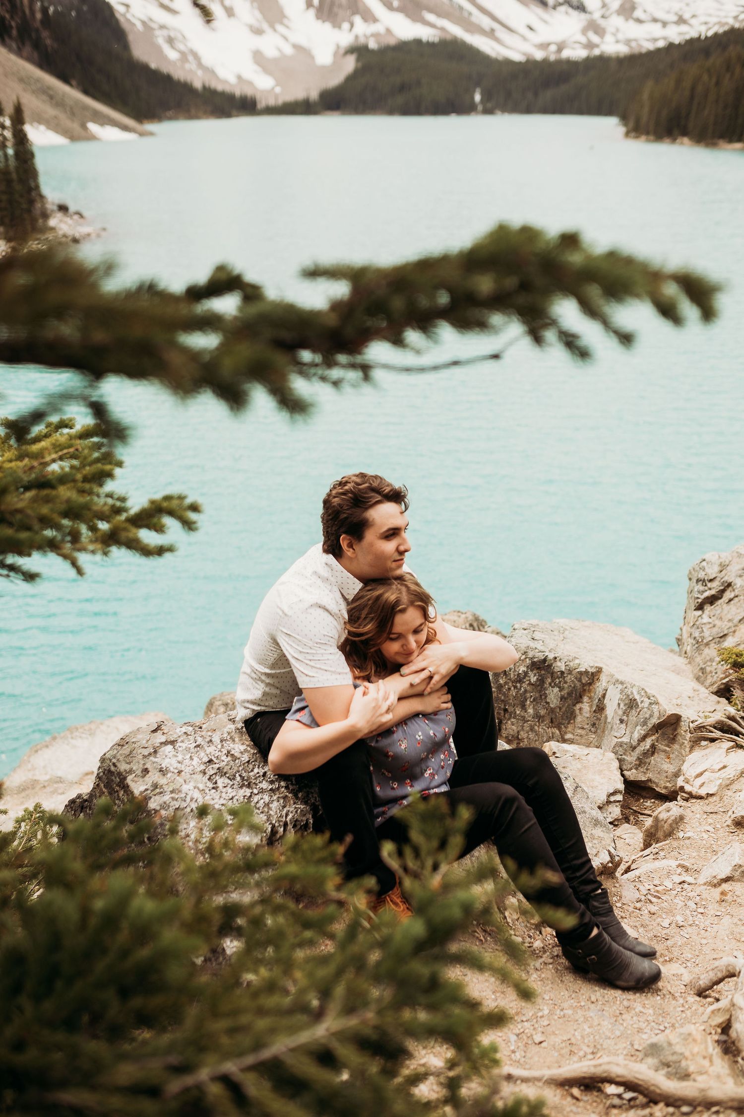 A tender moment shared on rocky outcrop overlooking turquoise alpine lake and mountain vista.