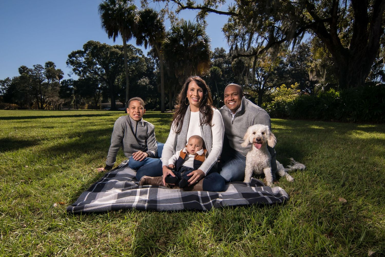 A family and their white dog enjoy a picnic on a blanket in a grassy field with trees behind them.