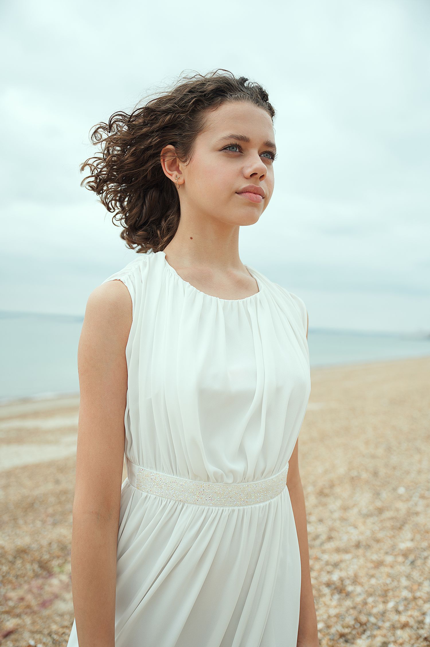 Woman in flowing white dress stands on pebble beach with windswept curly hair against cloudy sky.