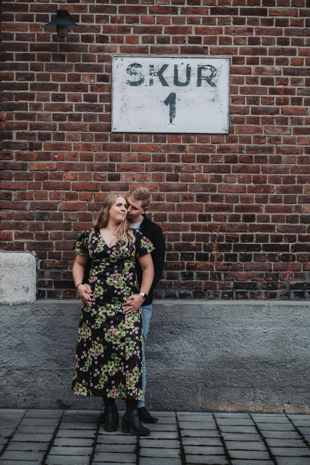 A couple poses against a brick wall with a 'SKUR 1' sign in an urban setting.