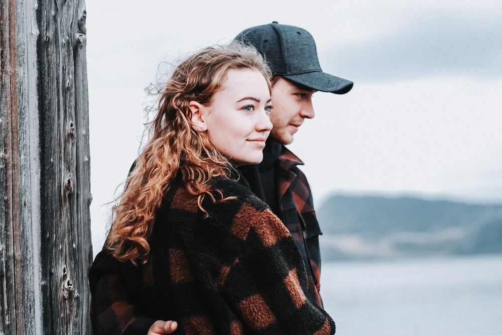 A romantic coastal portrait of a couple in plaid jackets overlooking a scenic waterway.