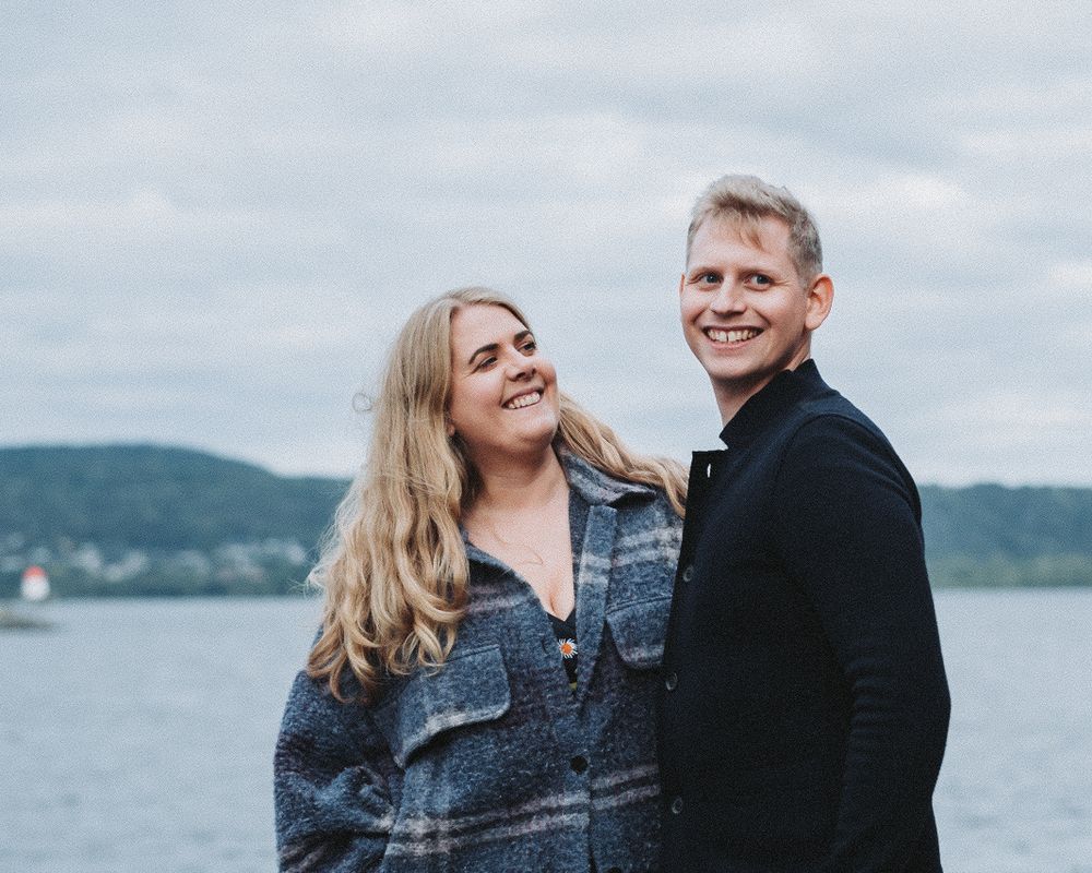 A couple in woolen jackets shares a joyful moment along a scenic waterfront vista.