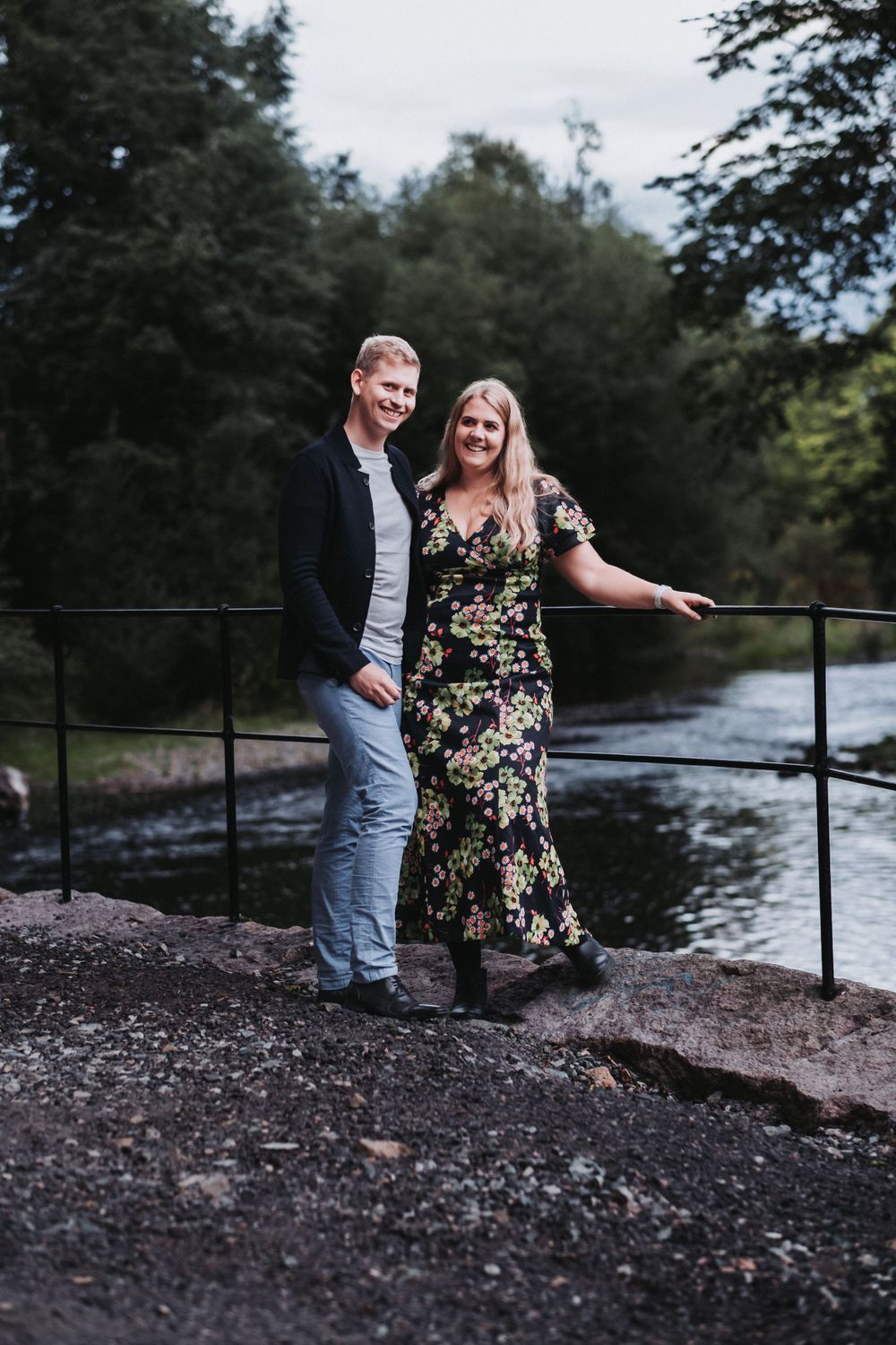 Couple posing on stone wall by river in countryside setting.