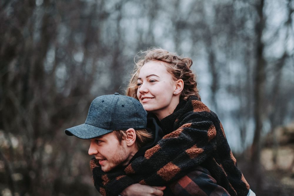 A lighthearted piggyback moment between a couple wearing coordinated plaid outfits in the woods.