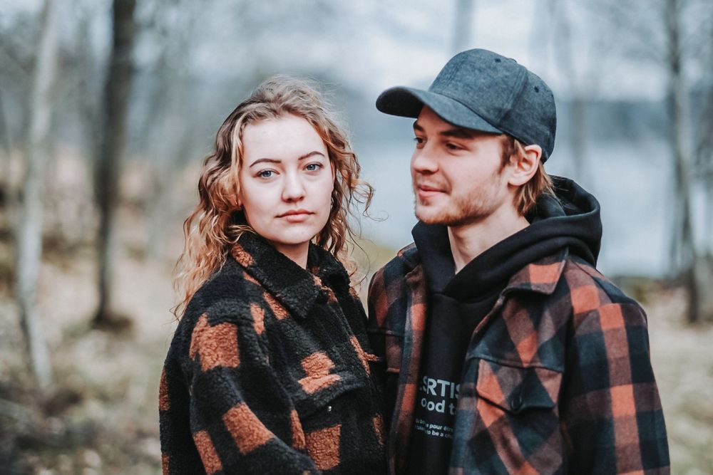 A couple in matching buffalo plaid jackets poses in an intimate portrait against a forest backdrop.