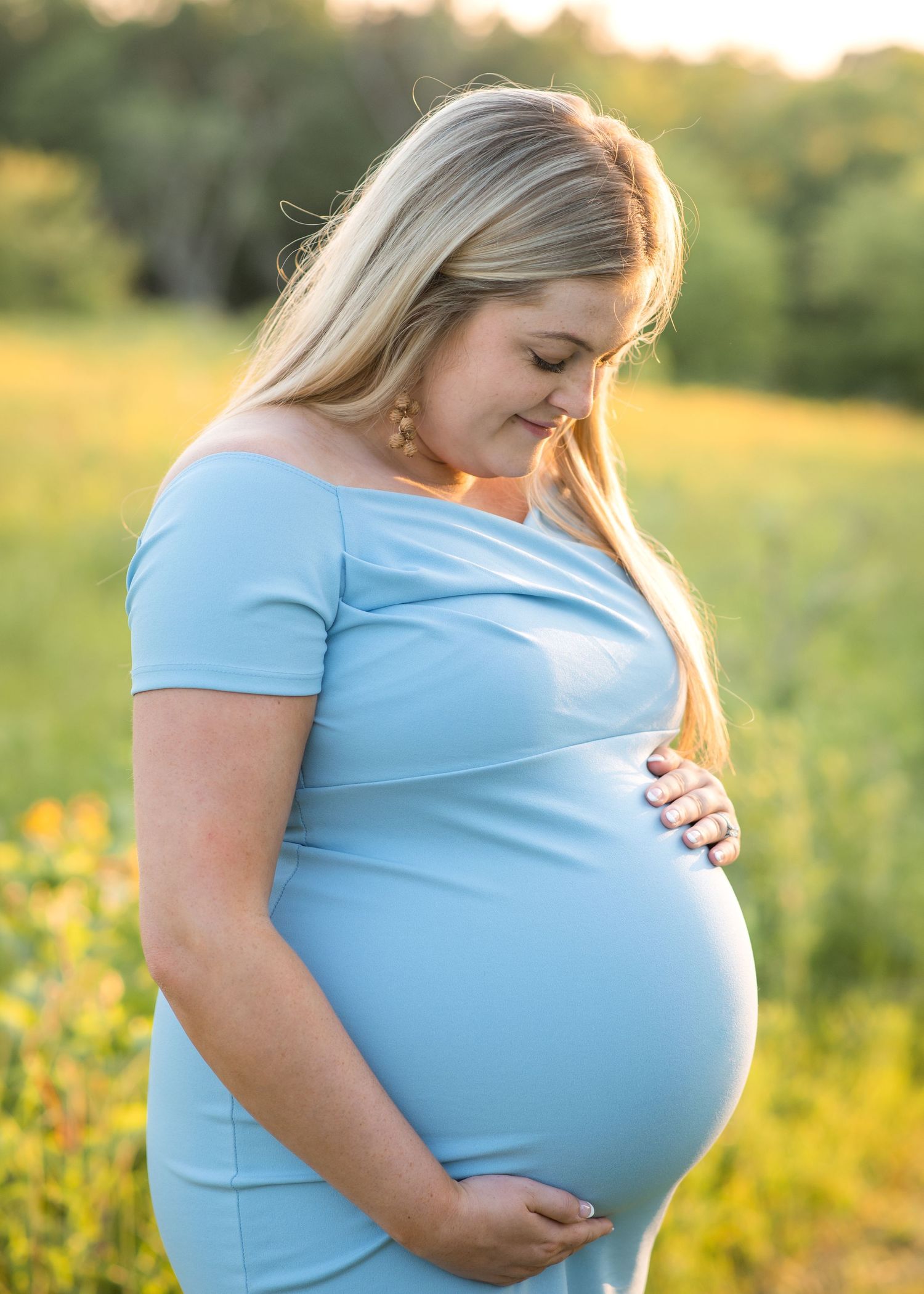Expecting mother in blue dress caresses baby bump during golden hour in wildflower field.