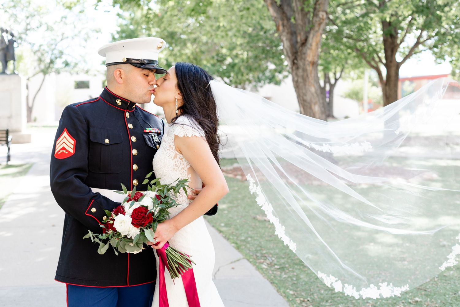 Military wedding portrait with flowing veil and red bouquet in outdoor garden setting.