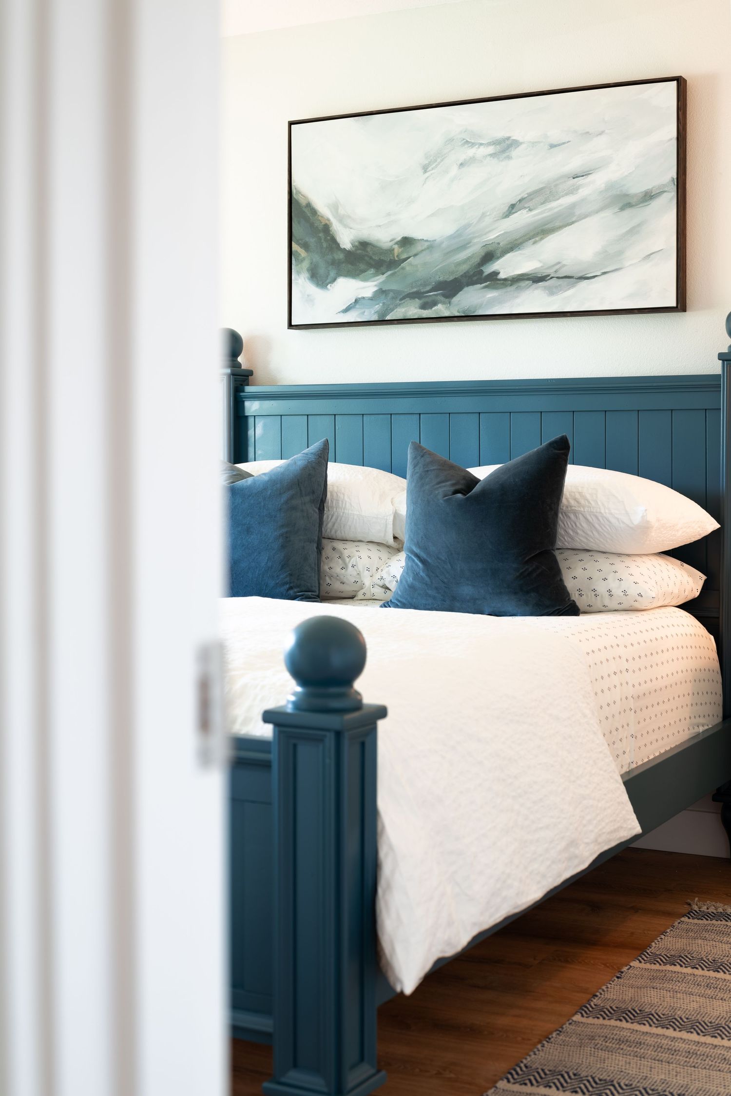 Modern bedroom interior with blue painted bed frame, abstract art, and white bedding.