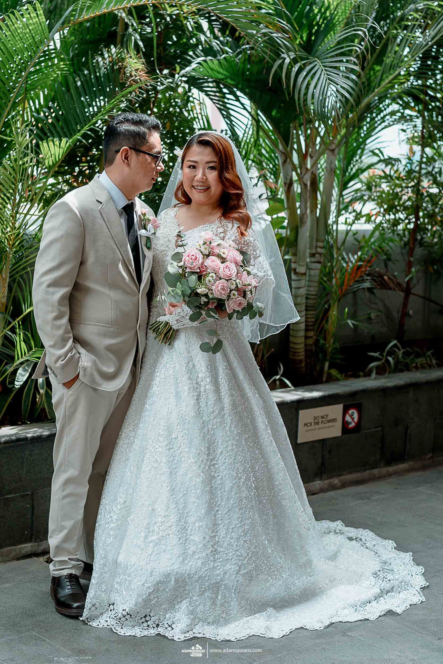 Wedding portrait with bride in lace gown and groom in beige suit against tropical greenery.