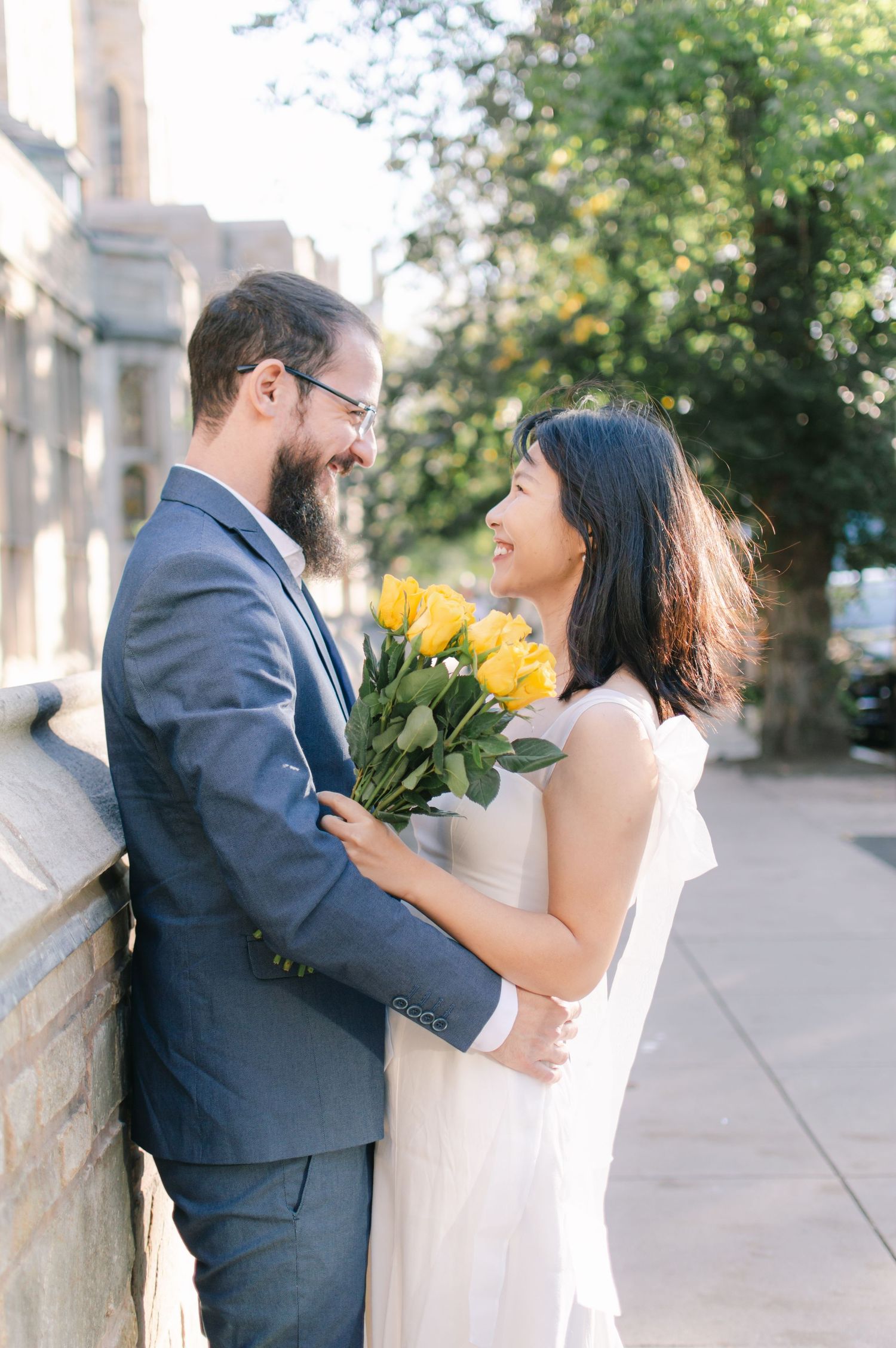 A bearded man in a blue suit and woman in white dress share a tender moment holding yellow roses outdoors.