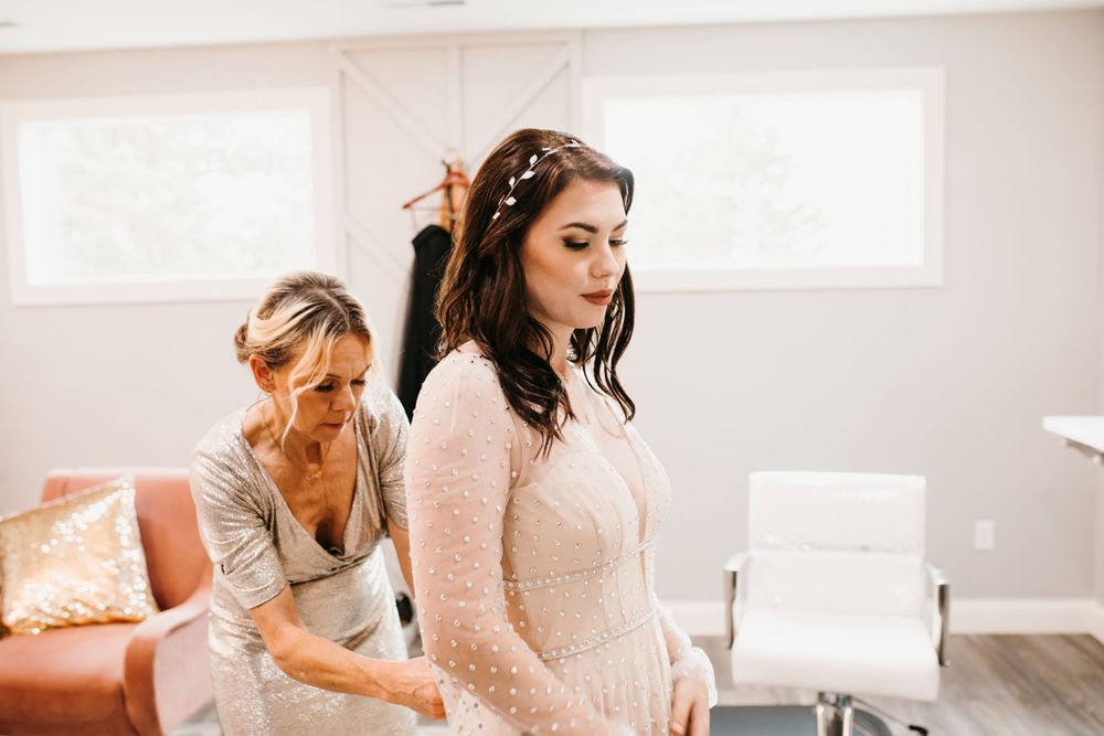 Mother helps bride adjust her pearl-embellished wedding gown in bright modern bridal suite before ceremony.