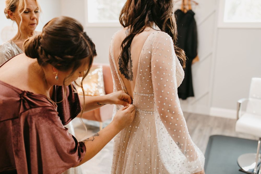 Bridesmaid in dusty rose off-shoulder dress carefully buttons back of bride's pearl-dotted wedding gown.