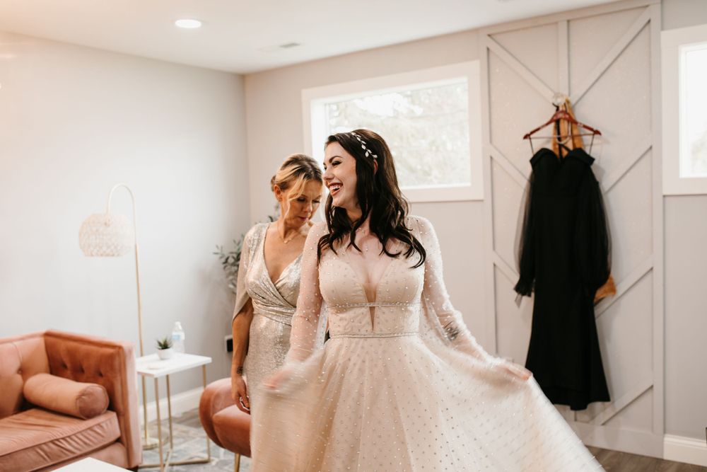 Bride laughs joyfully in sparkling long-sleeve wedding dress while mother smiles proudly in bright suite.