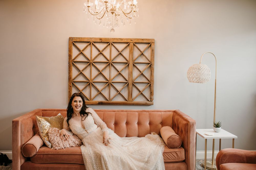 Radiant bride in lace wedding gown laughs while seated on pink velvet sofa under chandelier.