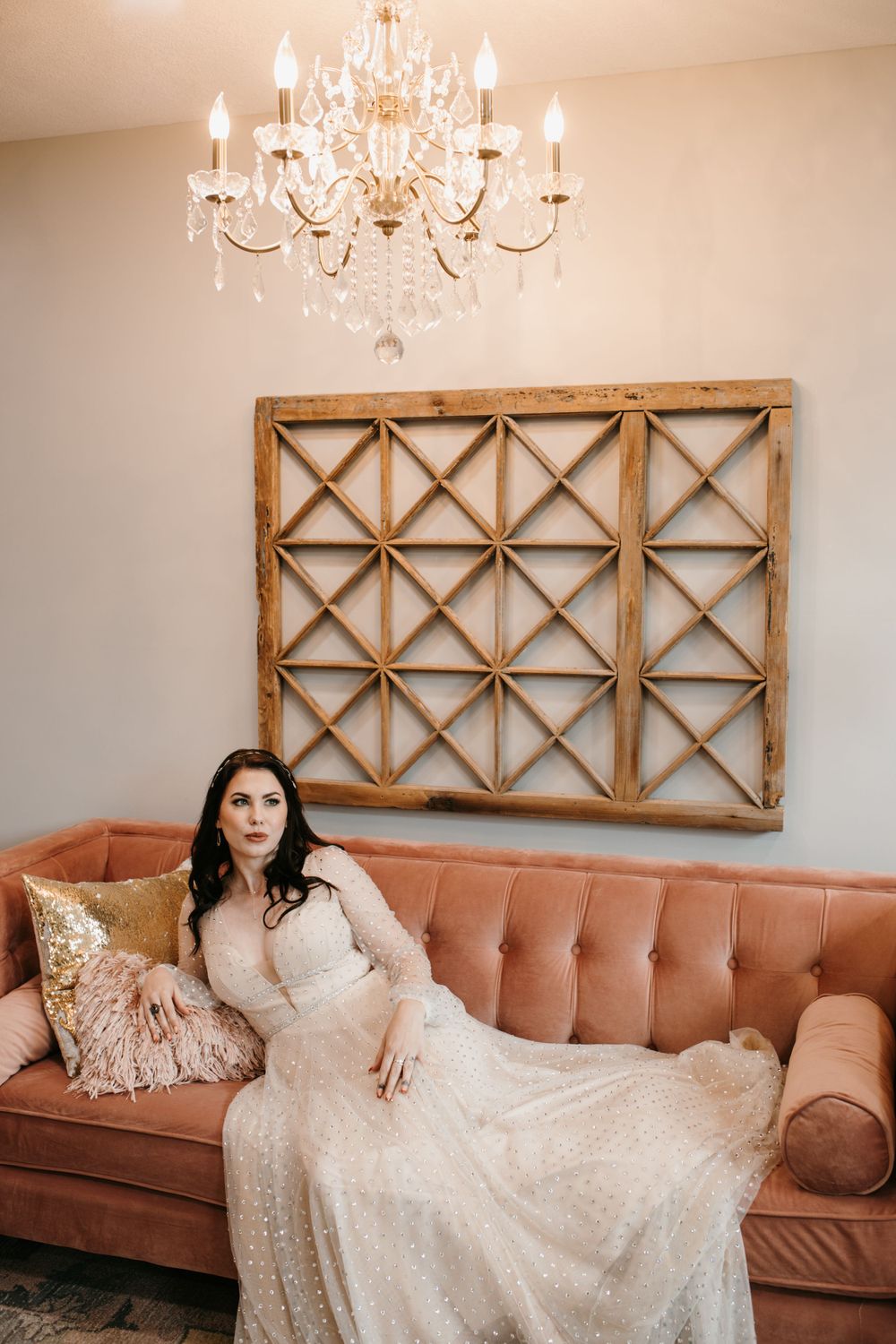 Elegant bride in beaded gown reclines on pink tufted sofa beneath wooden wall art and chandelier.