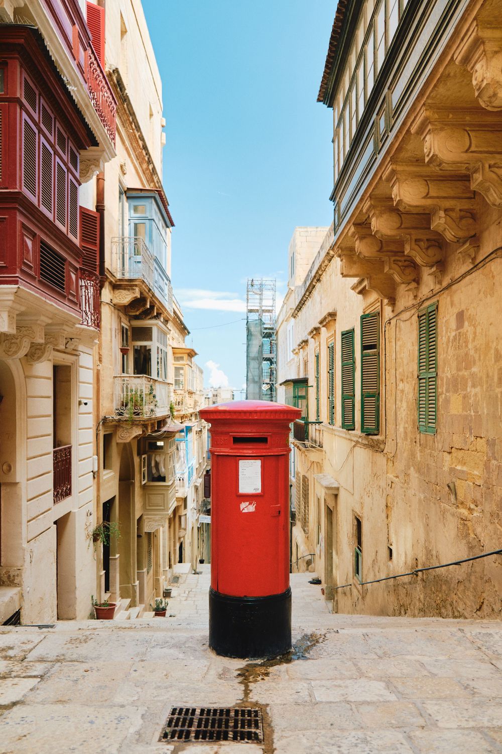 Red British post box stands in a narrow traditional Maltese street with limestone buildings.