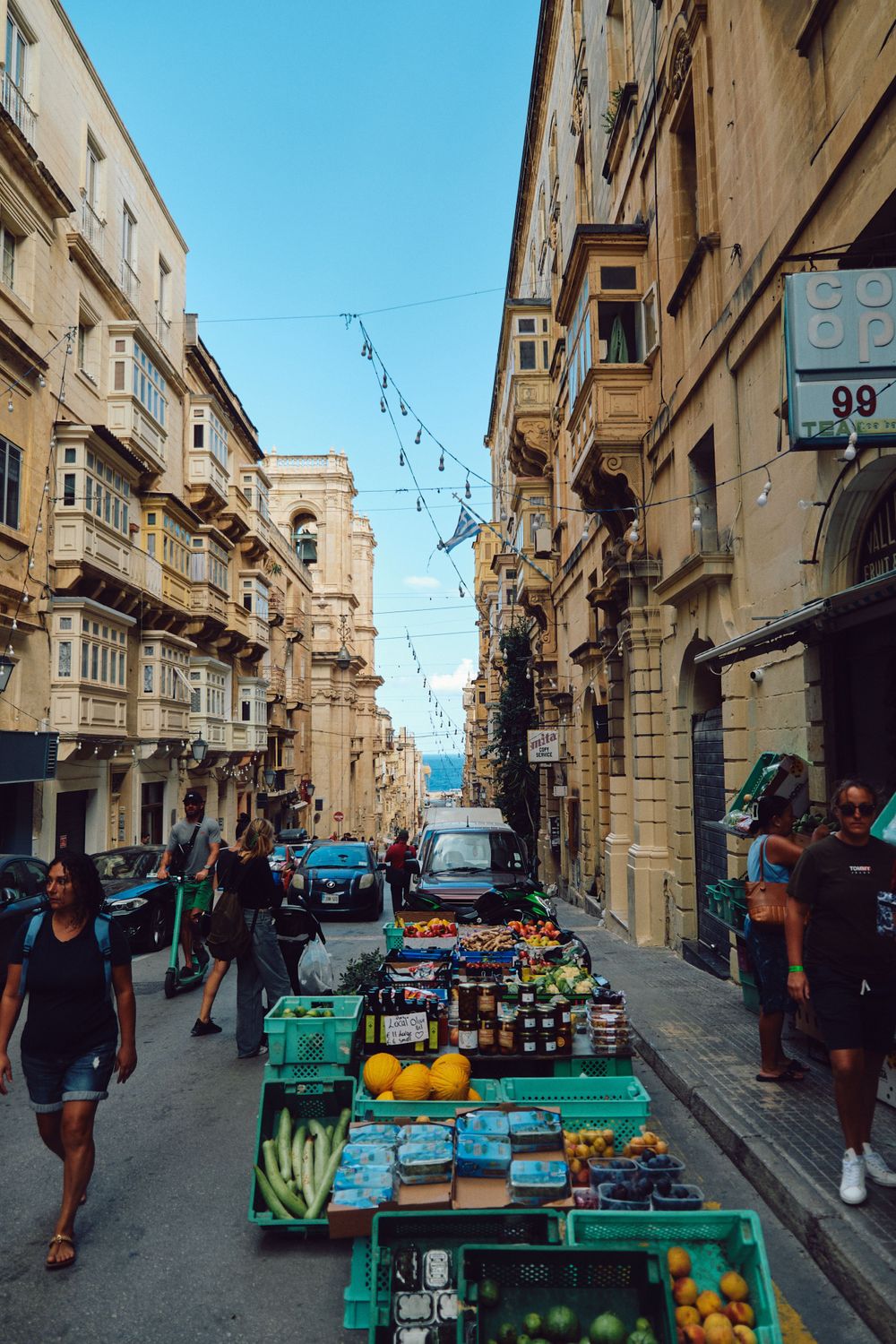 Narrow street market in Malta with vendors selling fresh produce and goods between historic limestone buildings.