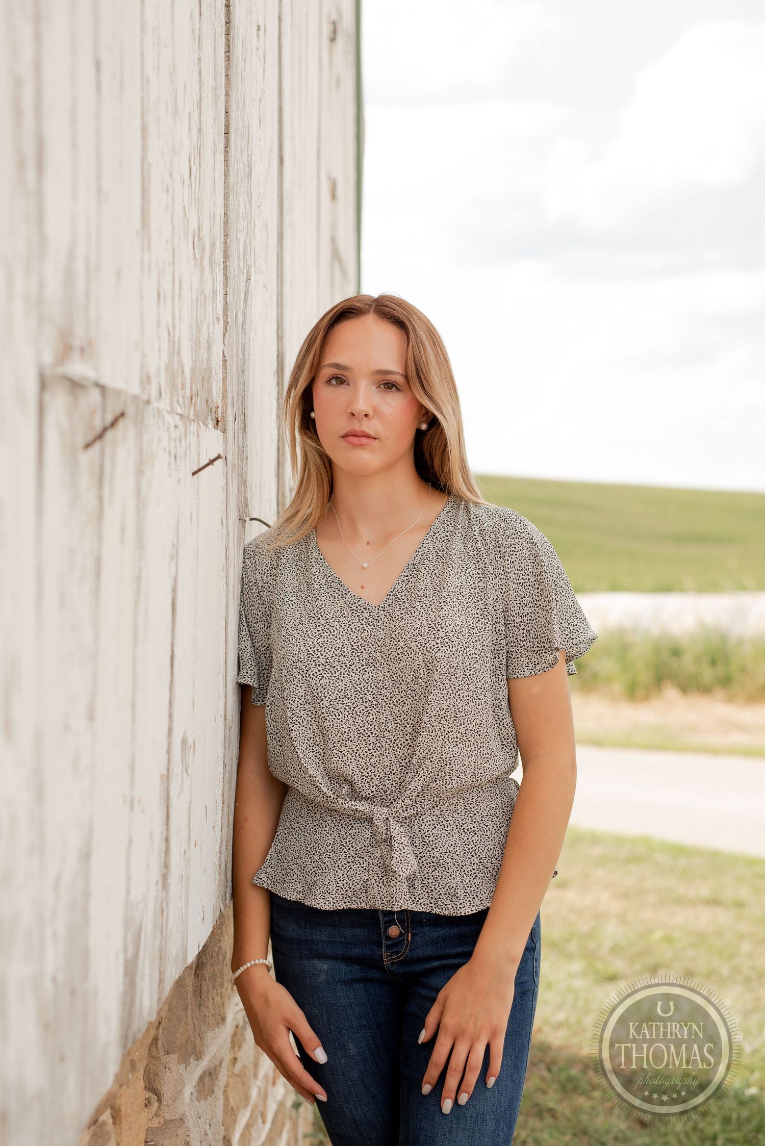 A person in a v-neck printed top stands against a white barn wall with farmland in the background.