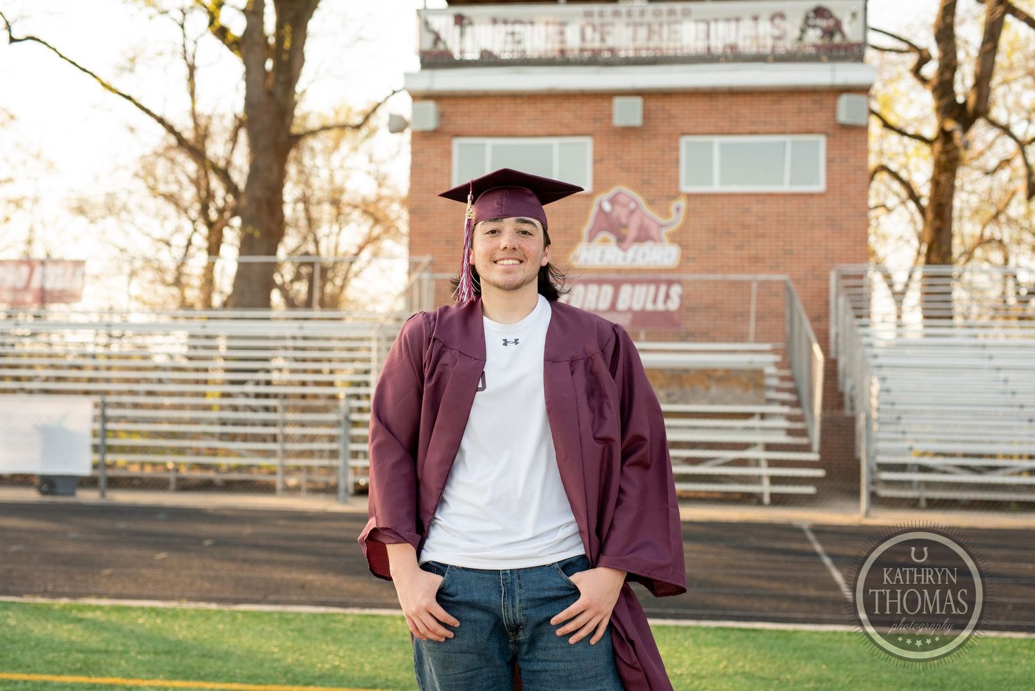 Graduate in maroon gown poses confidently in front of stadium bleachers during sunset photo shoot.