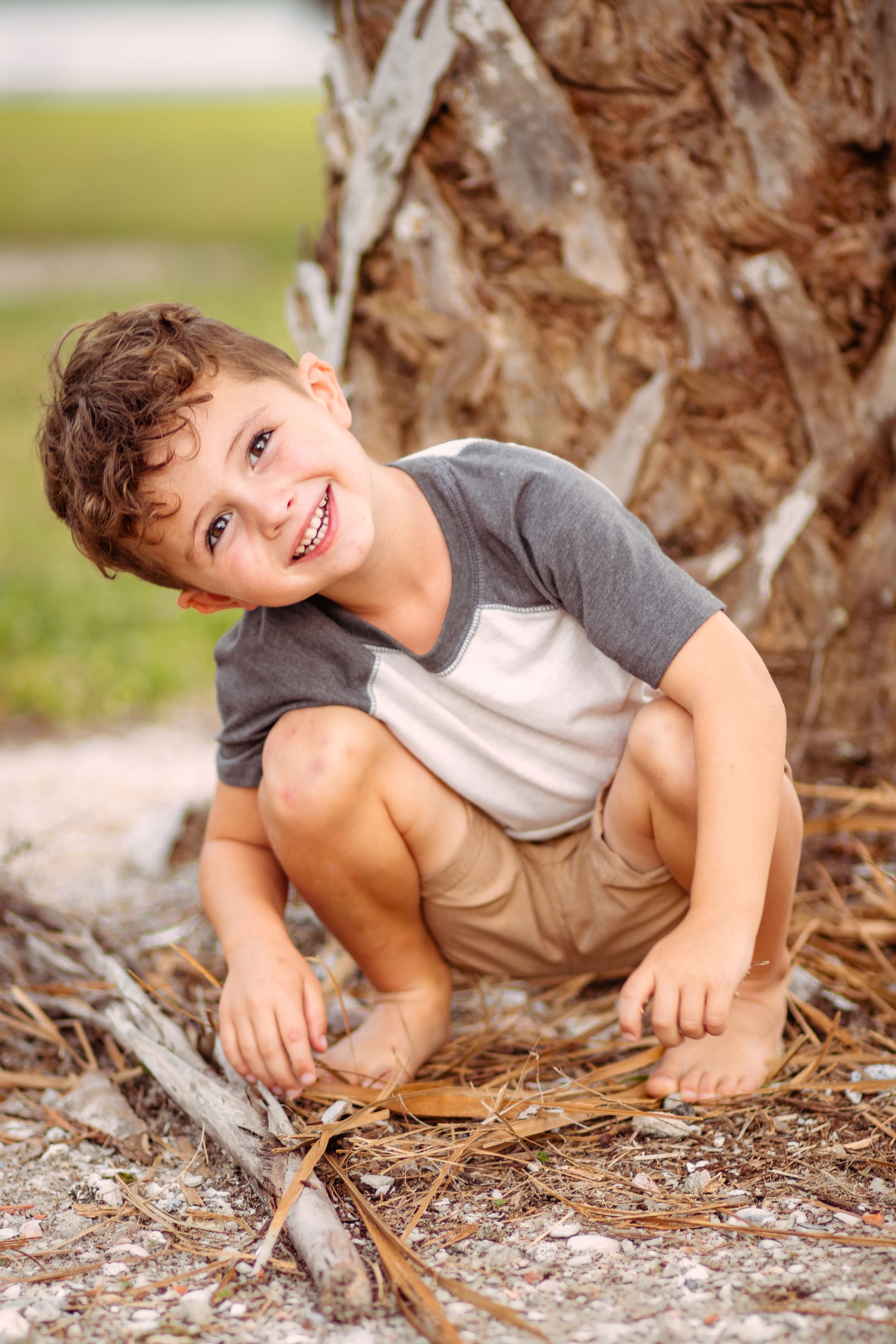 Smiling young boy crouching barefoot near a palm tree, playing outdoors on a sandy ground with dry leaves.