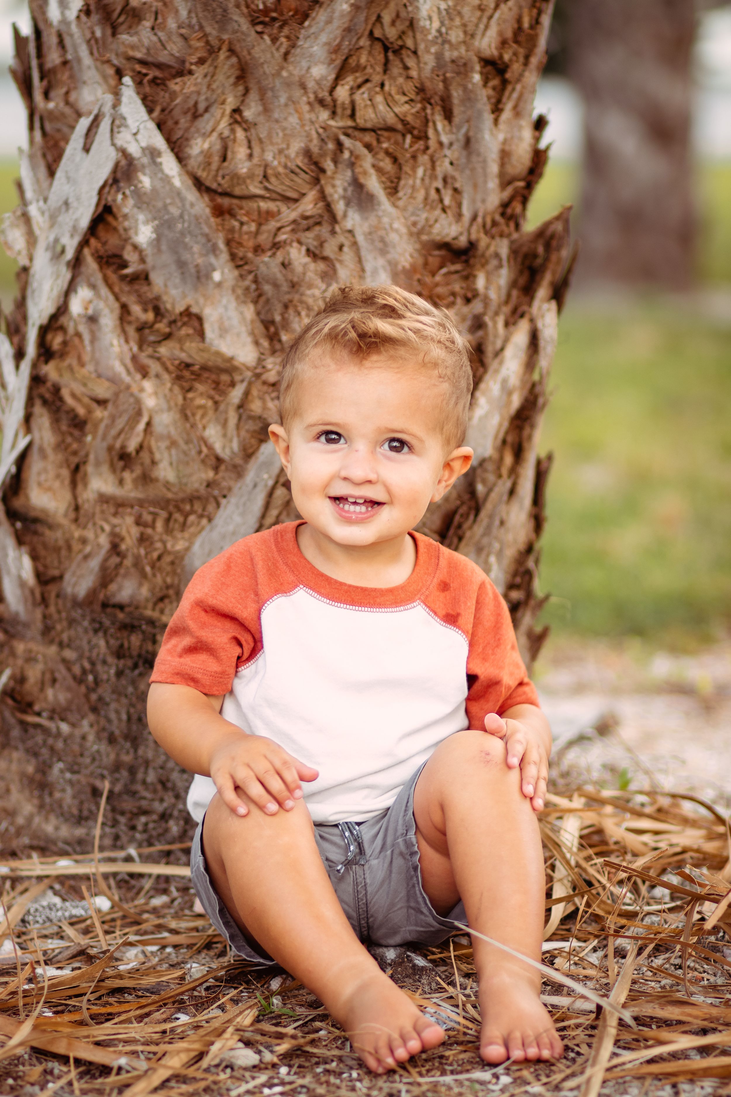 Smiling toddler boy in orange and white shirt sitting barefoot by a palm tree on dry grass outdoors.