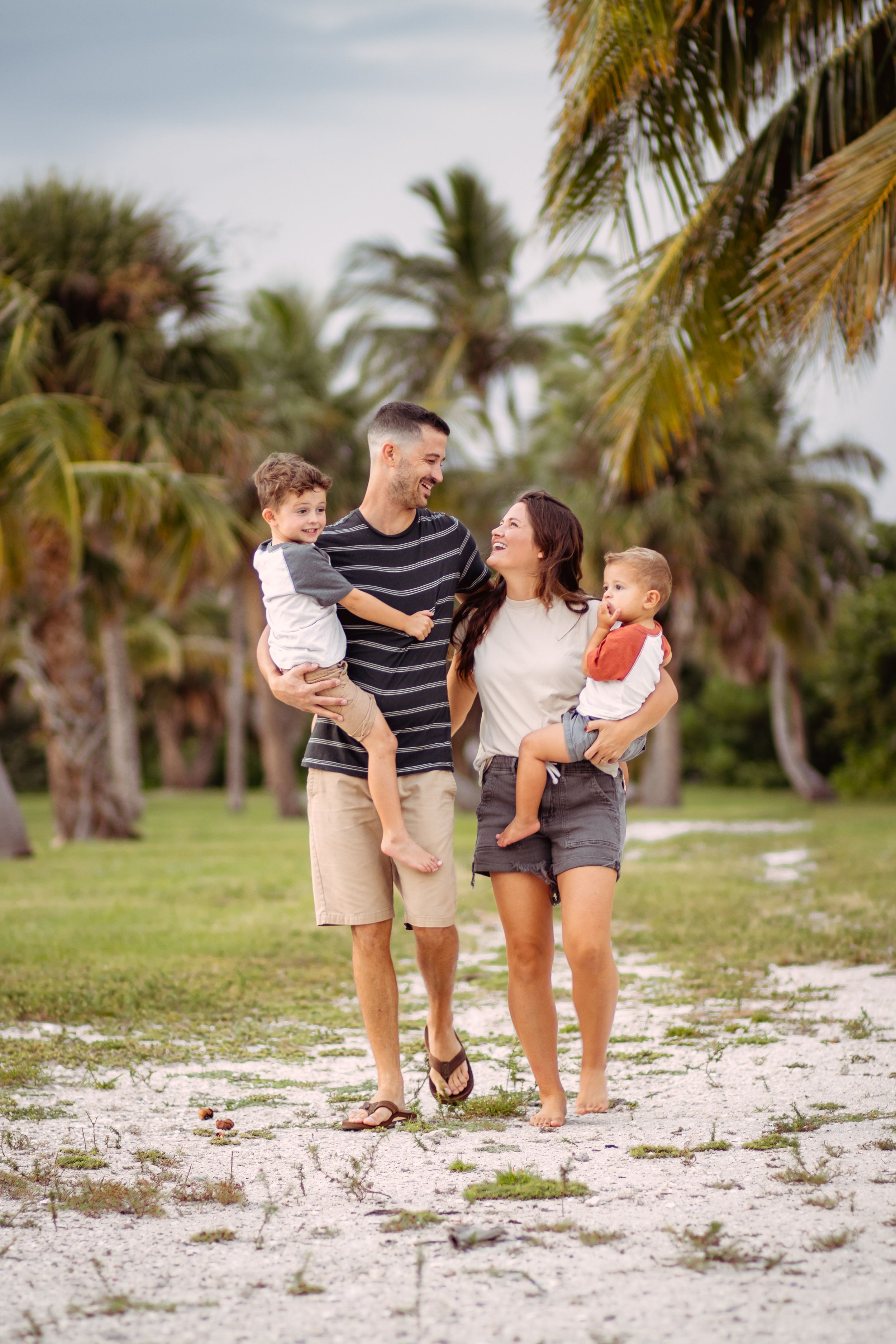 Happy family of four posing outdoors among palm trees on a sandy beach, parents holding two young toddlers.