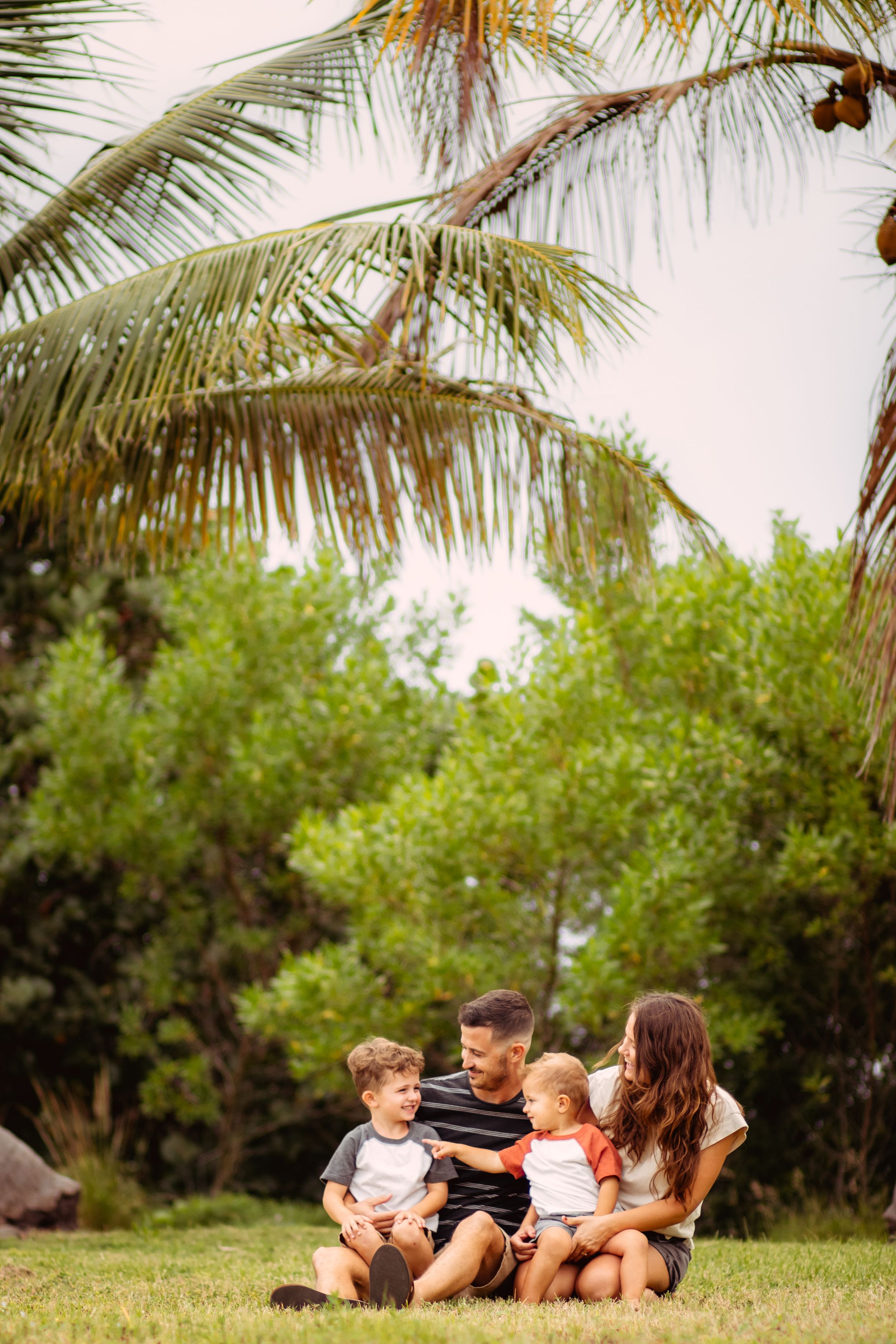Family of four sitting on grass under palm trees in a tropical garden, parents playing with two young children.