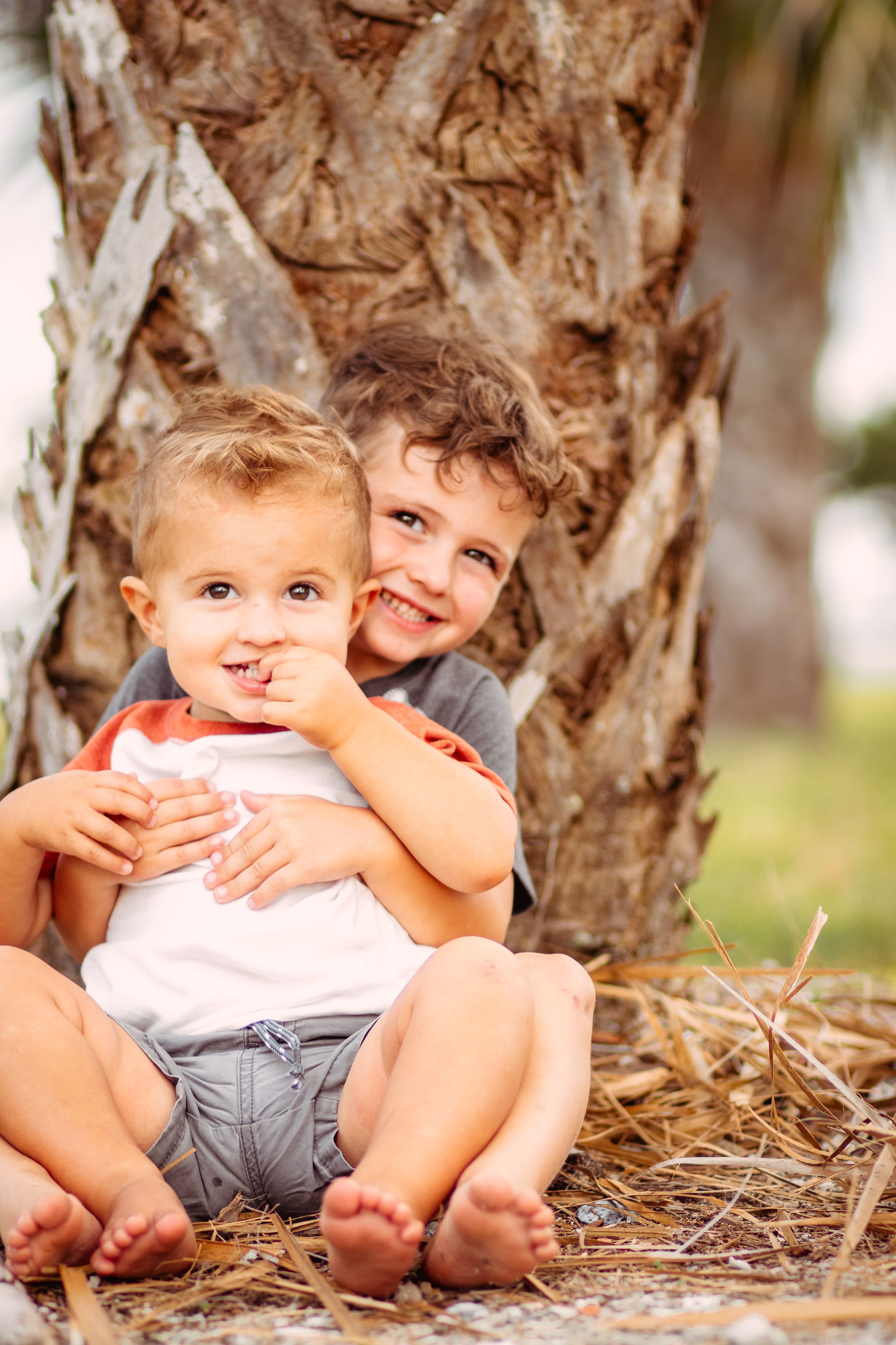 Two young boys sitting barefoot by a palm tree, smiling and hugging outdoors on a sunny day.