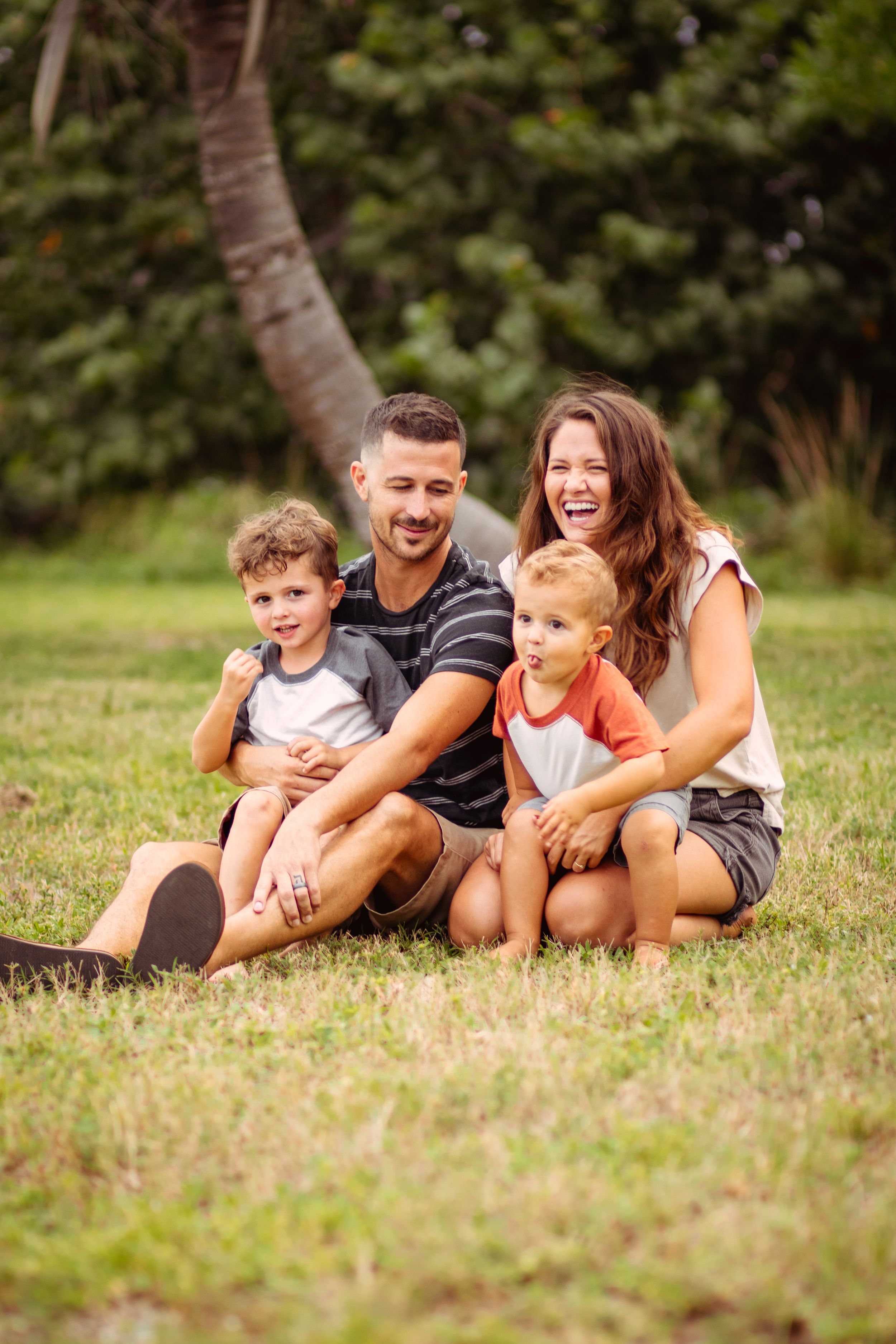 Happy family of four sitting on green grass outdoors, parents laughing with two young boys in a park setting.