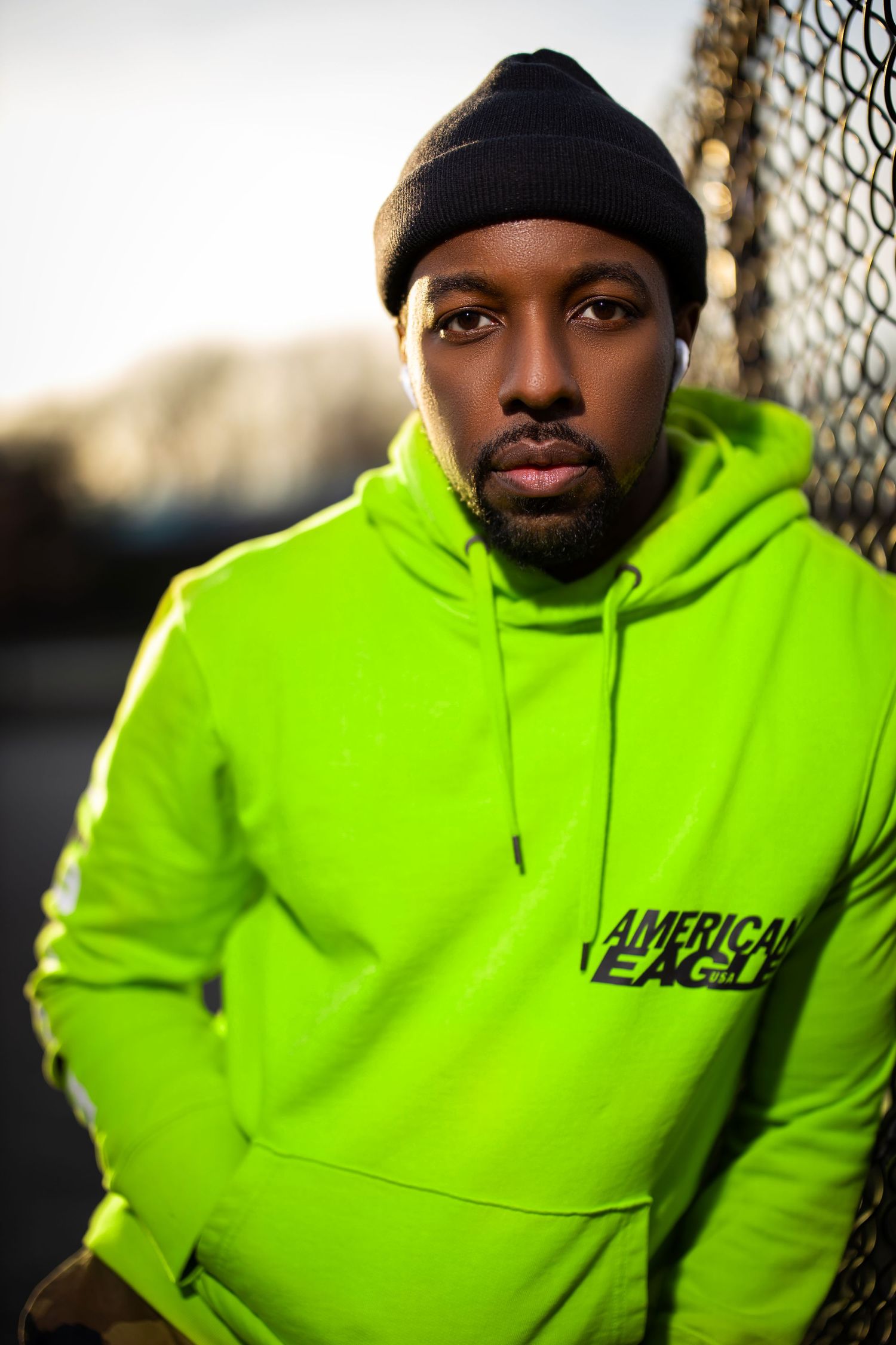 Portrait of person in neon yellow hoodie and black beanie against chain link fence.