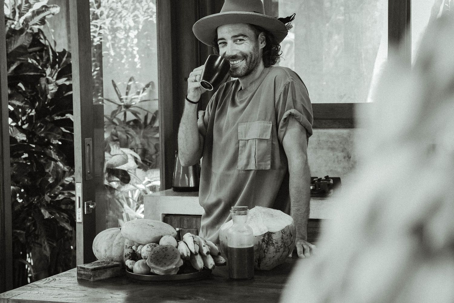 Black and white photo of person in hat with fruits on rustic wooden counter.