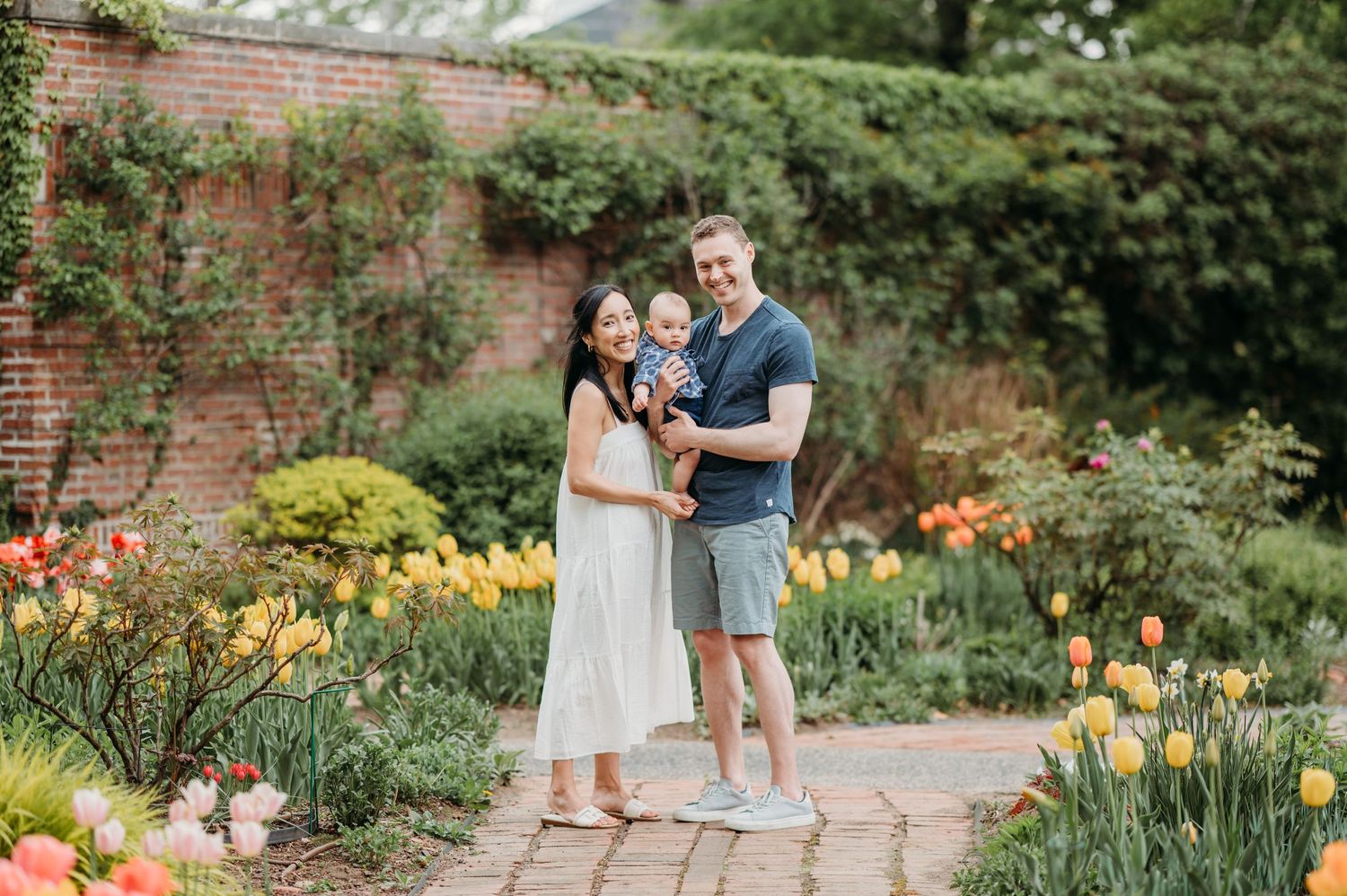A loving family poses together on a garden path surrounded by blooming tulips and an ivy wall backdrop.
