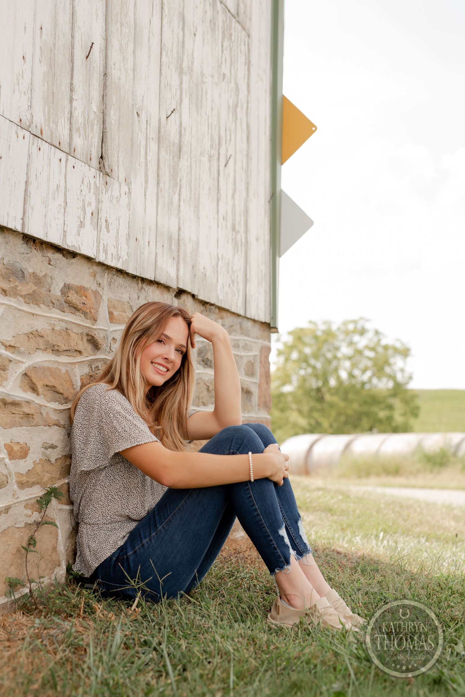 A person in a gray top and jeans relaxes against a rustic barn wall with rolling hills in the background.