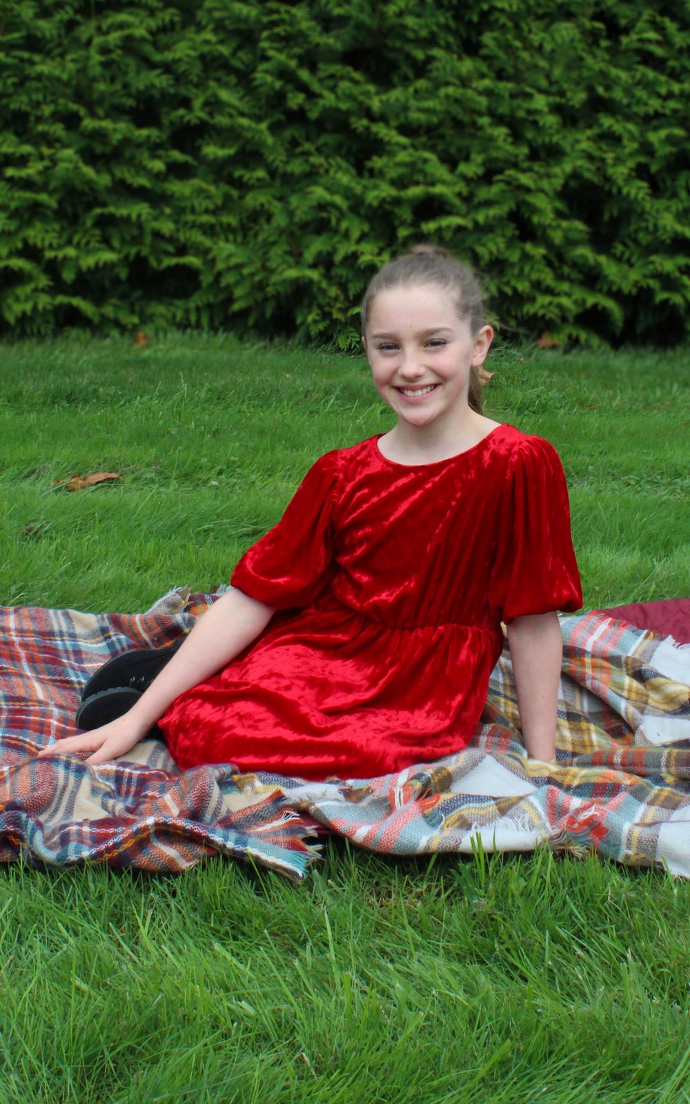 Person in red velvet dress sits smiling on plaid blanket on grass.
