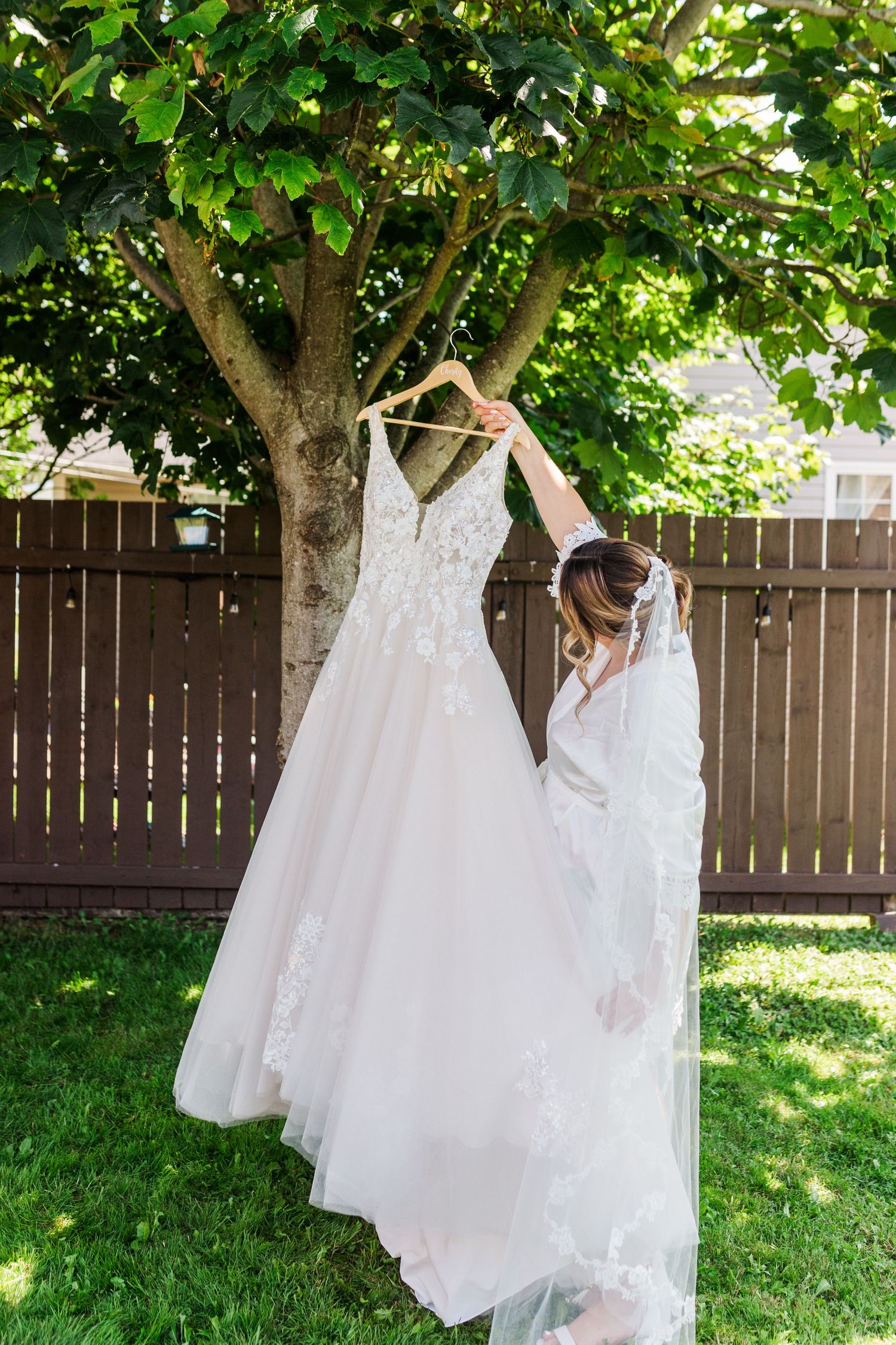 Beautiful white wedding dress hanging from a tree branch against a brown fence in a backyard garden setting.
