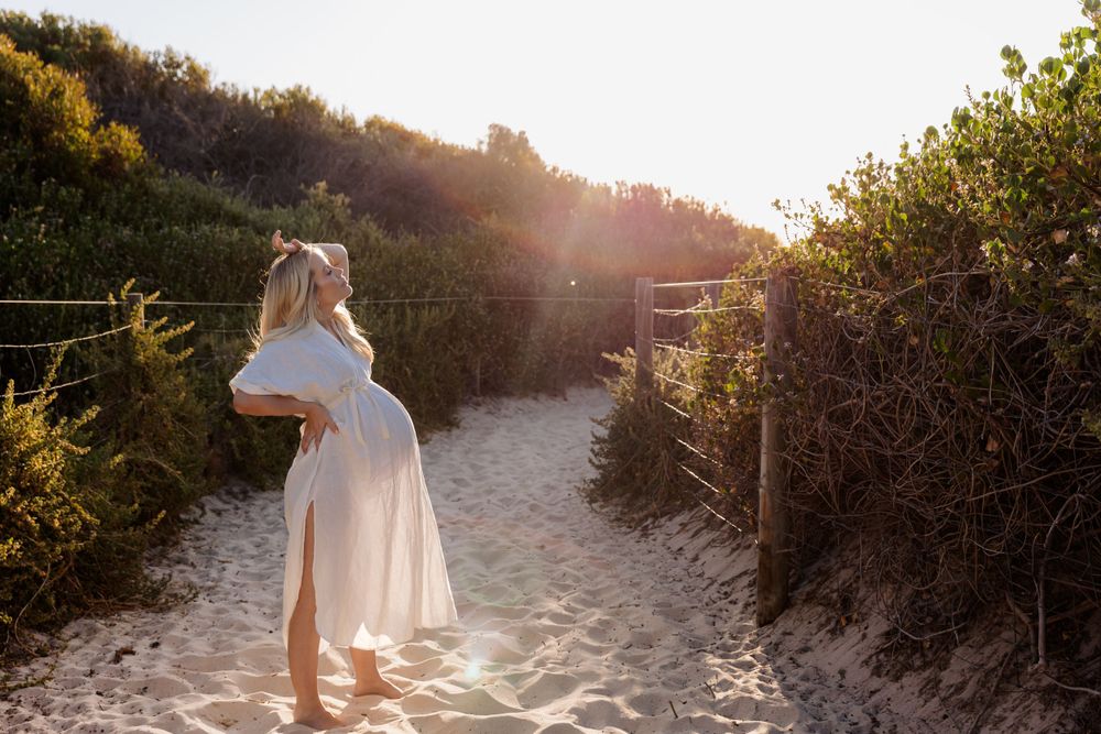 Silhouette of pregnant woman in flowing dress walking along beach path at sunset.