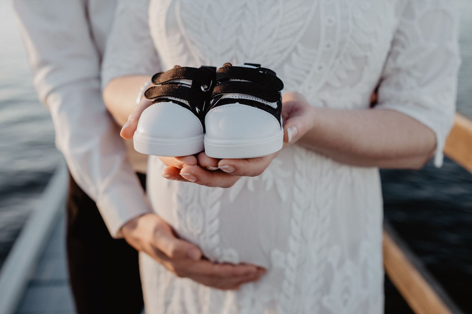 Close-up of hands holding small white baby shoes against lace dress background.