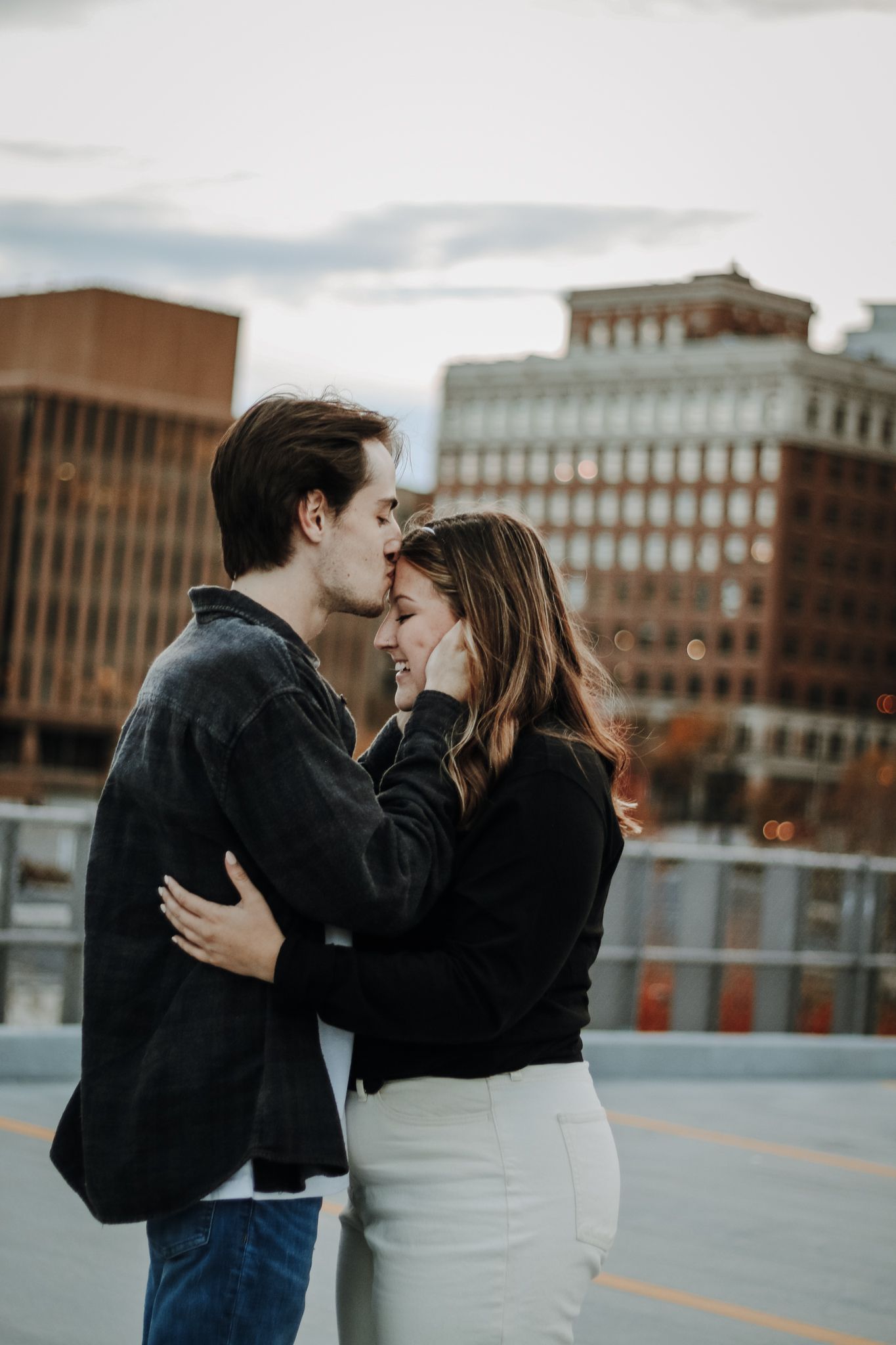 A romantic couple embraces on a rooftop parking deck with urban buildings in the background.