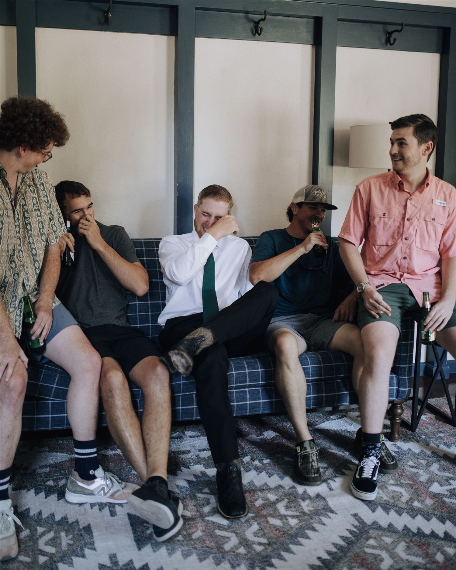 Groomsmen laugh together while sitting on a patterned couch in casual attire.