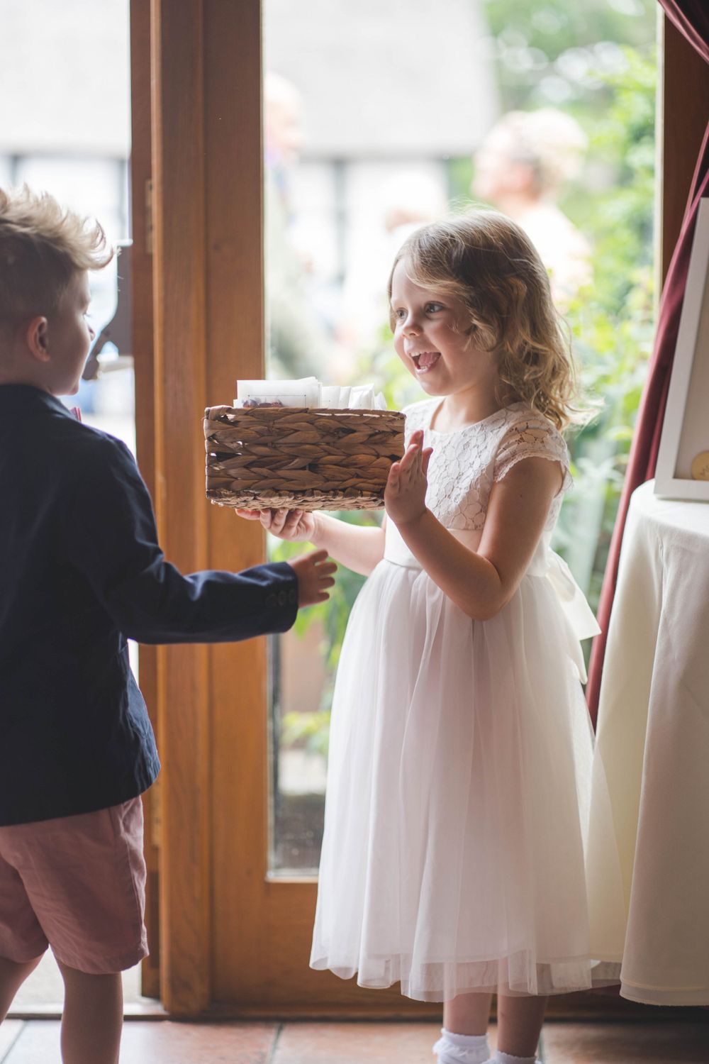 Young flower girls in white dresses play together near wooden windows.