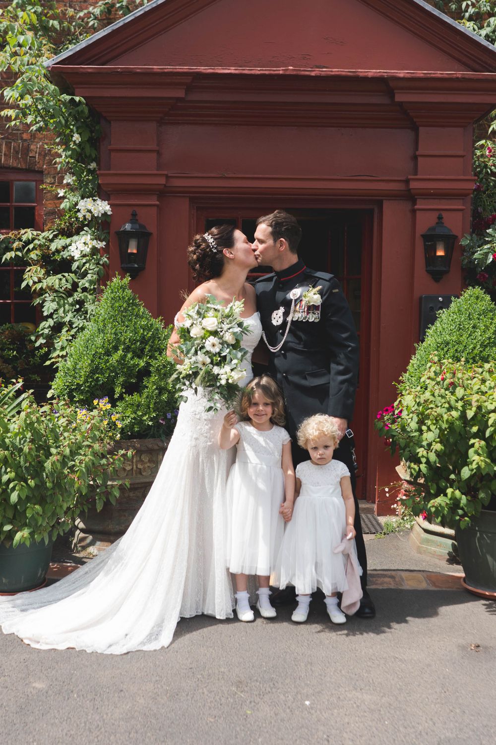 Wedding couple sharing kiss at red doorway with flower girls in white dresses.