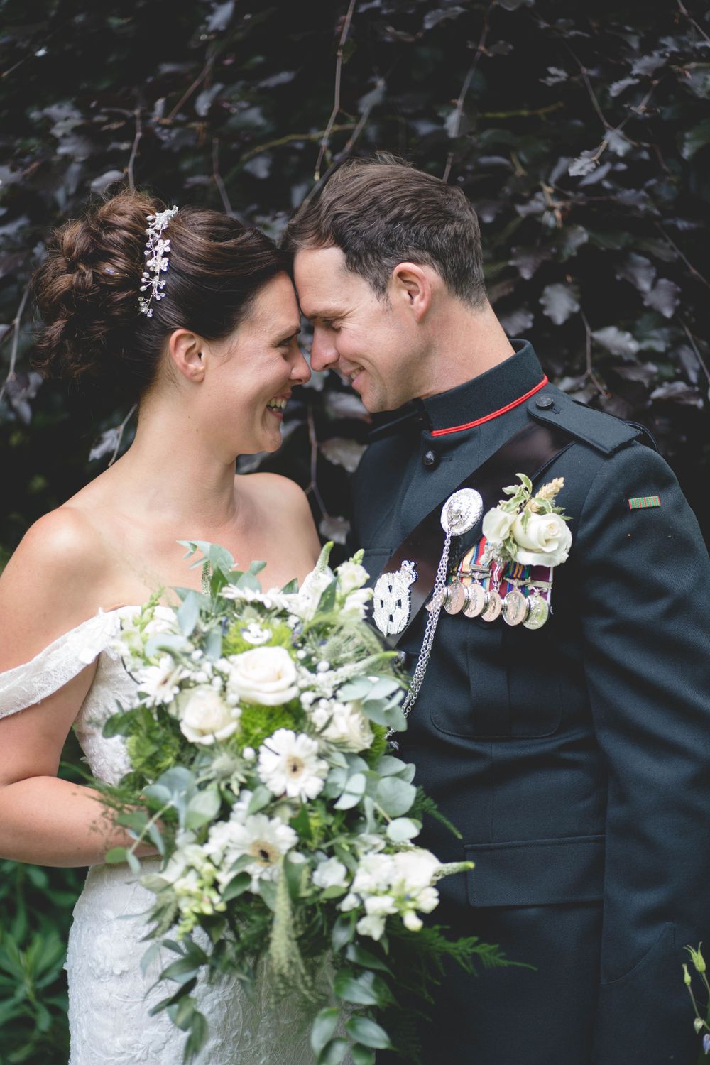 Military groom and bride share intimate moment against dark foliage backdrop.