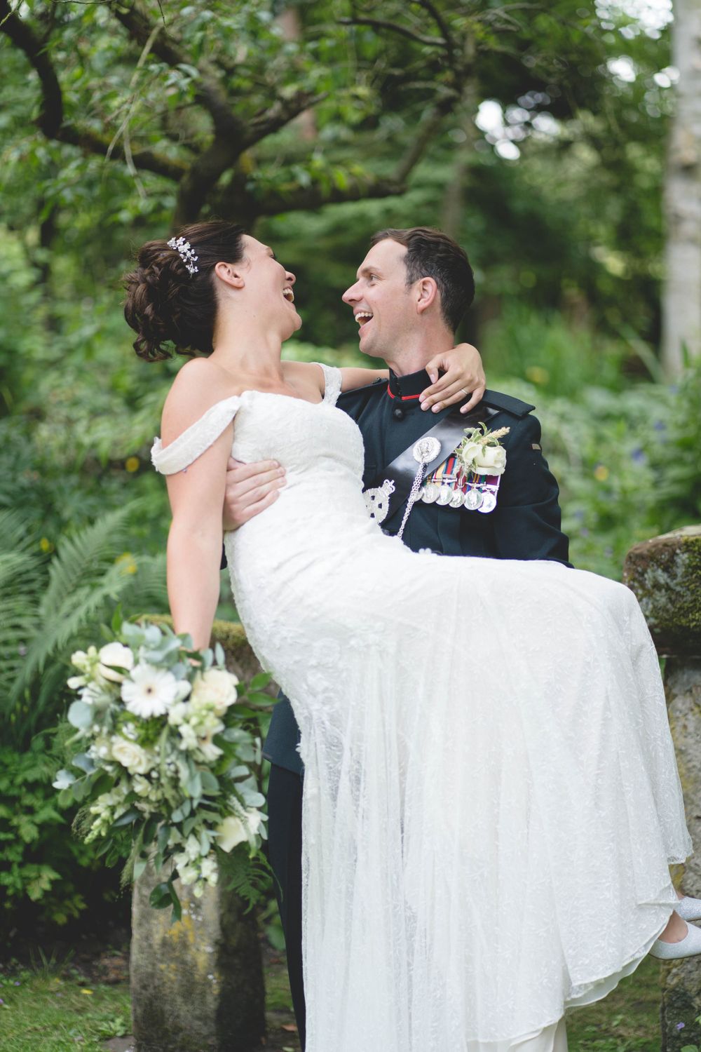 Military groom carries bride in white lace dress among garden greenery.