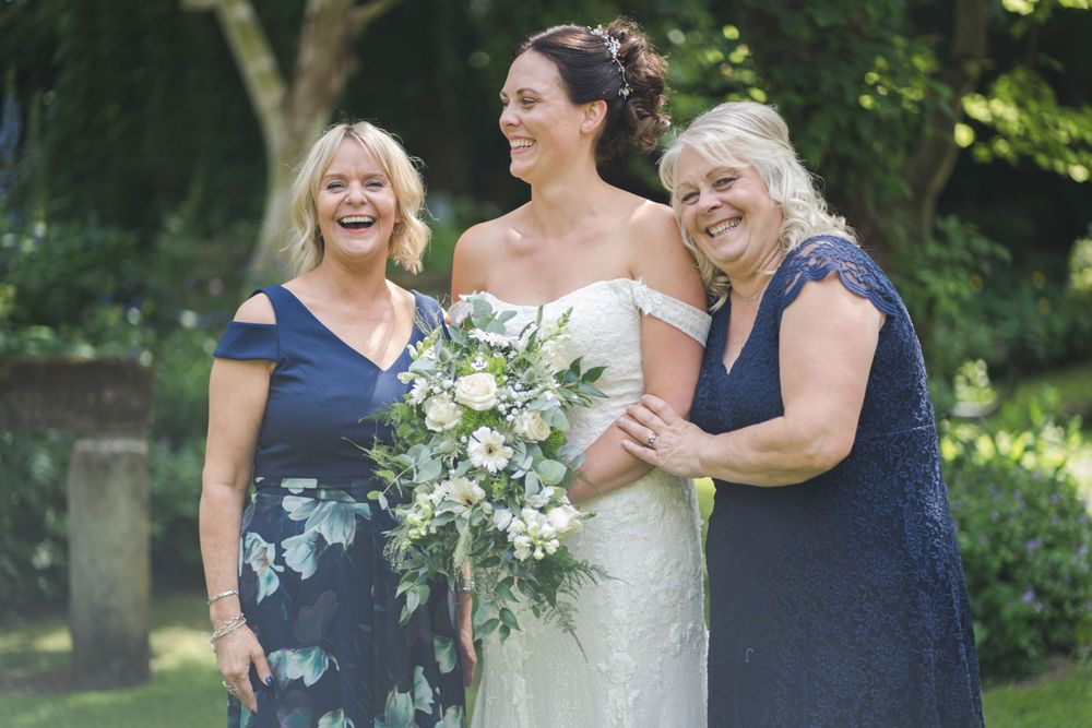 Wedding party poses together in natural outdoor garden setting with greenery and white floral bouquets.