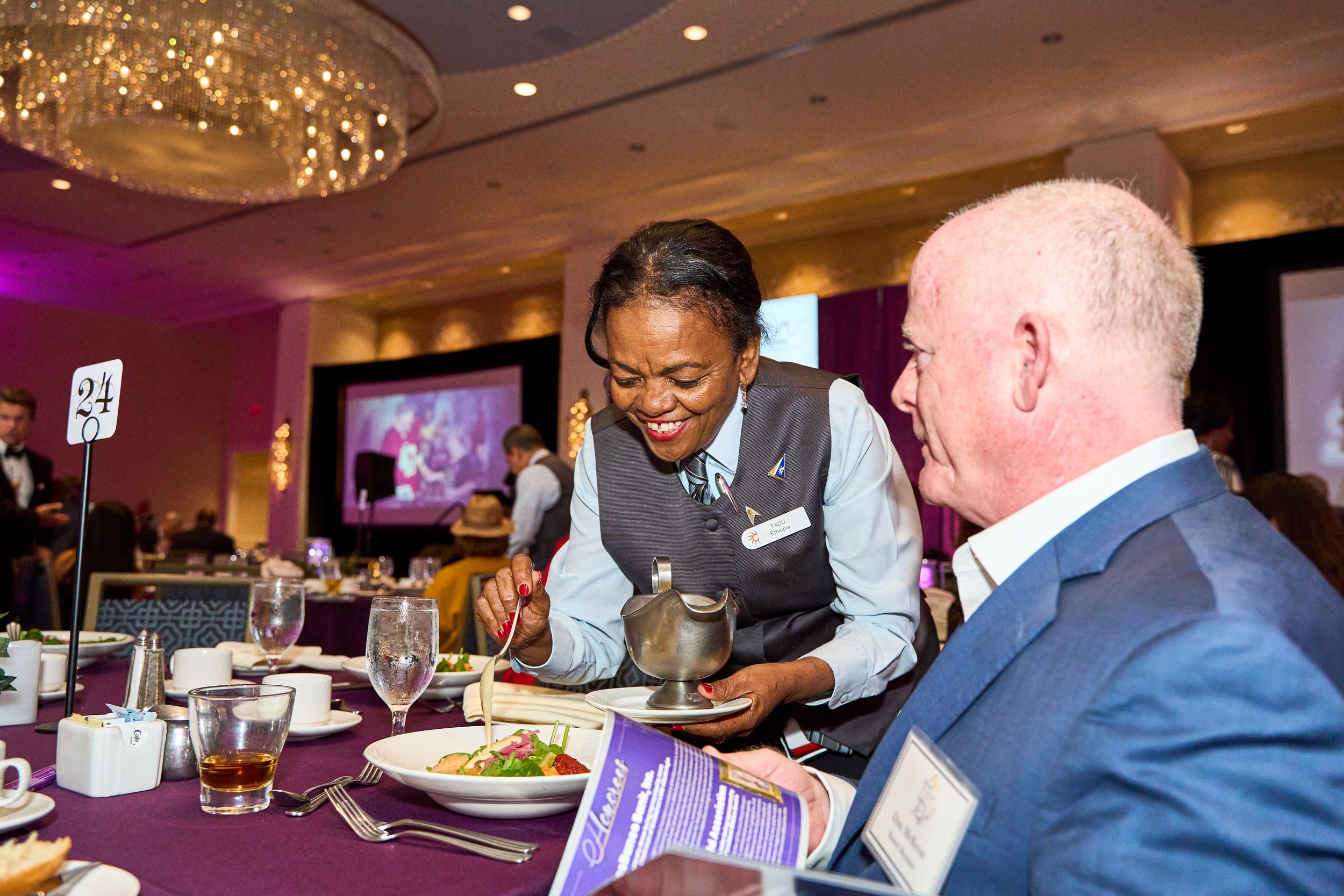 Server presenting a plated meal to guest at elegant dinner event with purple lighting.