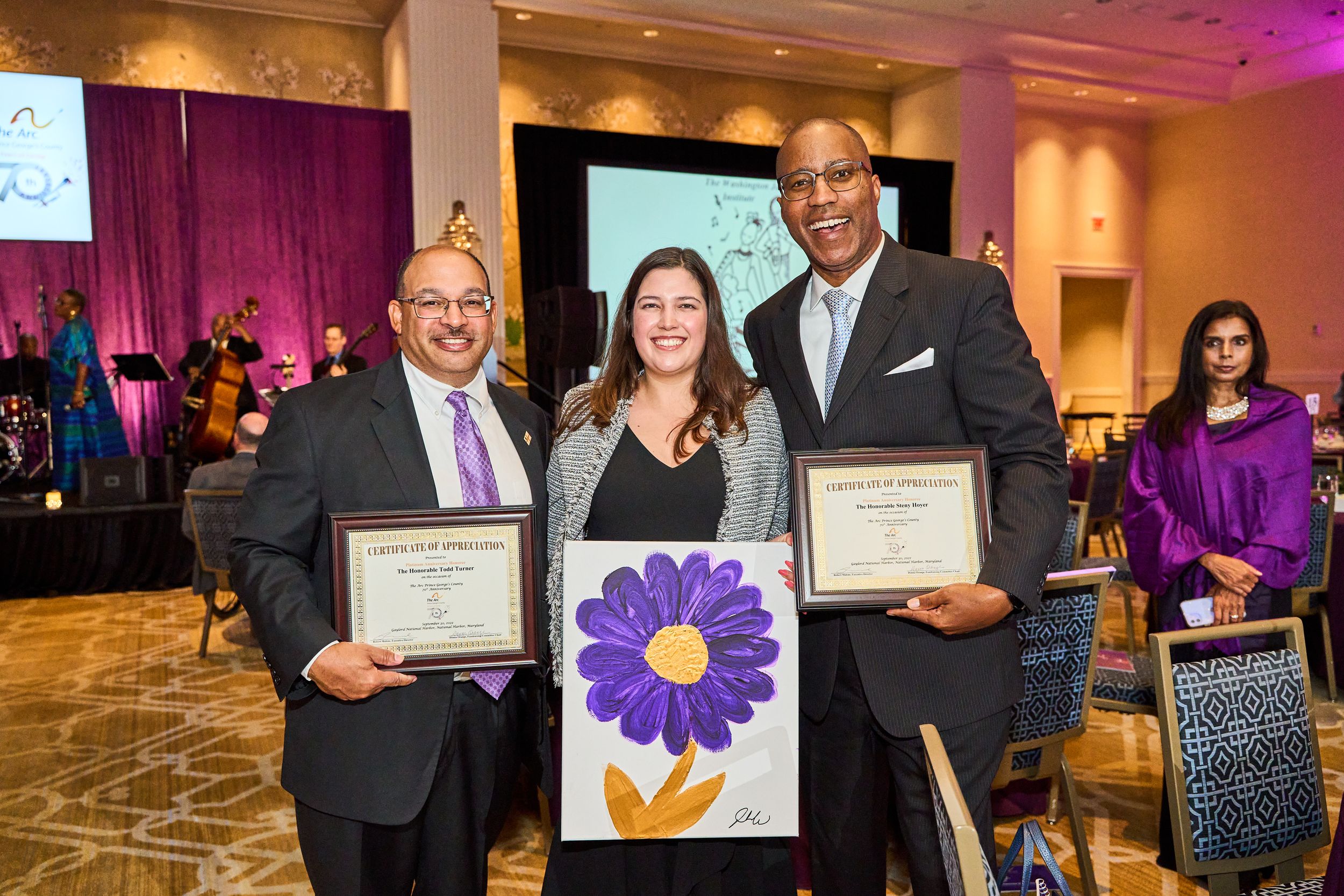 Three people displaying awards and artwork at formal recognition ceremony.