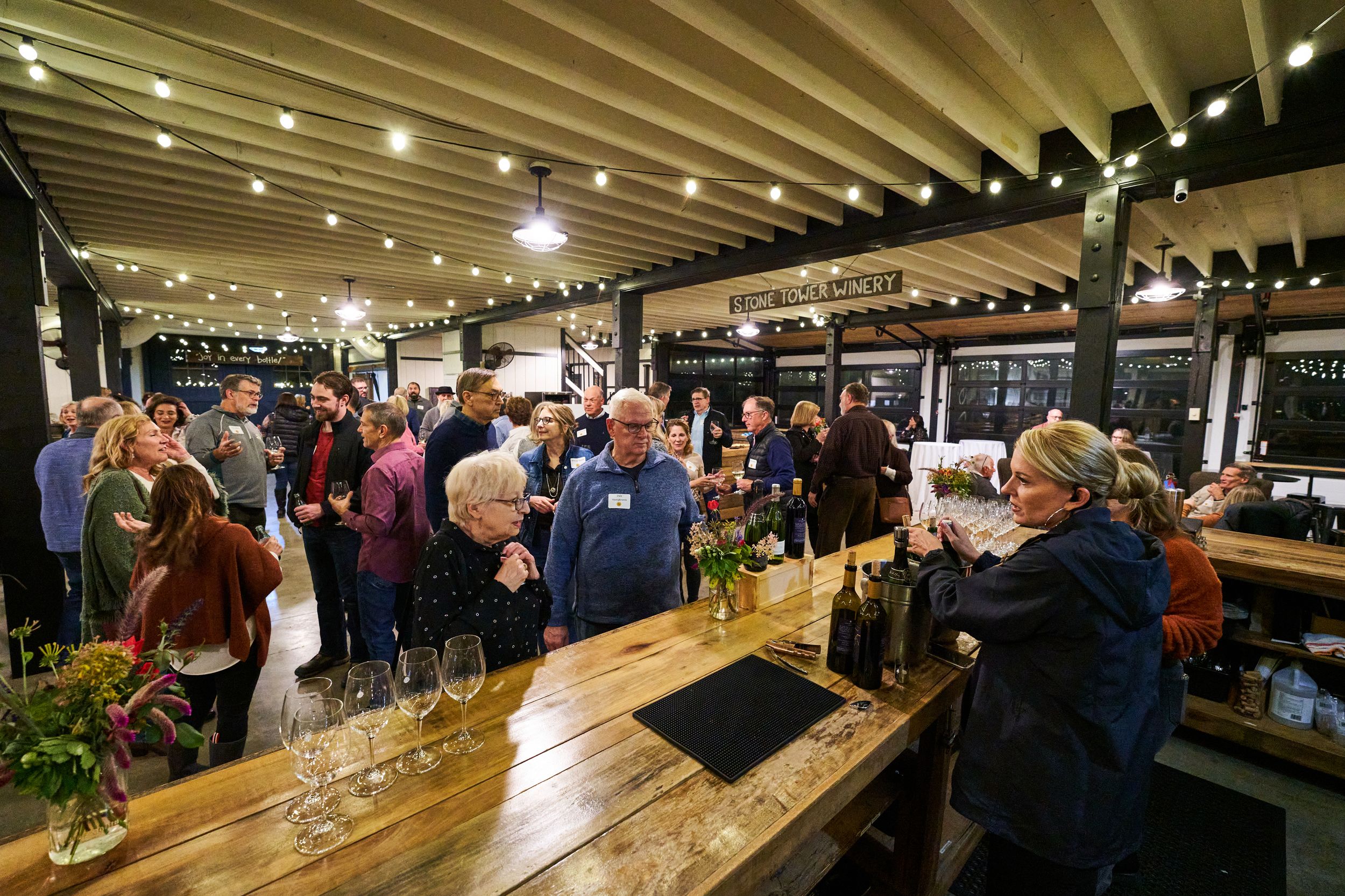Crowded bar event with wooden rustic interior and string lights overhead.