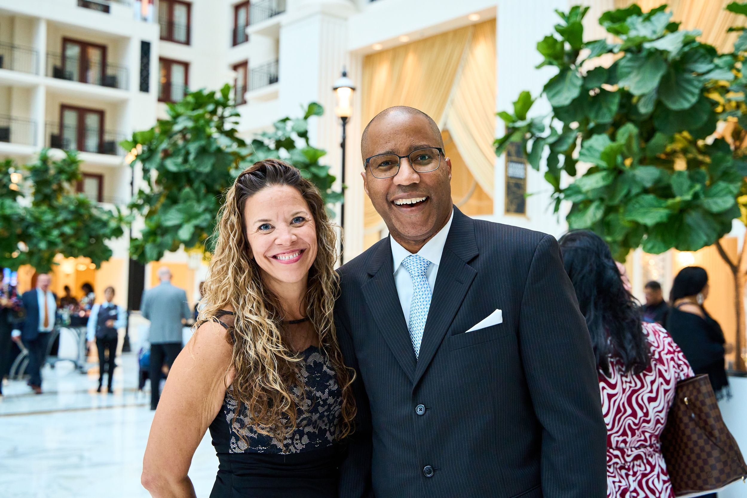 Two people posing together at an upscale social gathering with greenery in the background.